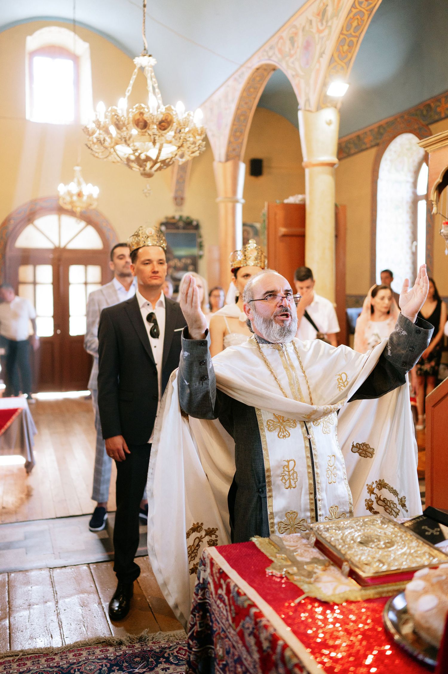 Priest performing religious ceremony with raised hands in decorated Orthodox church.