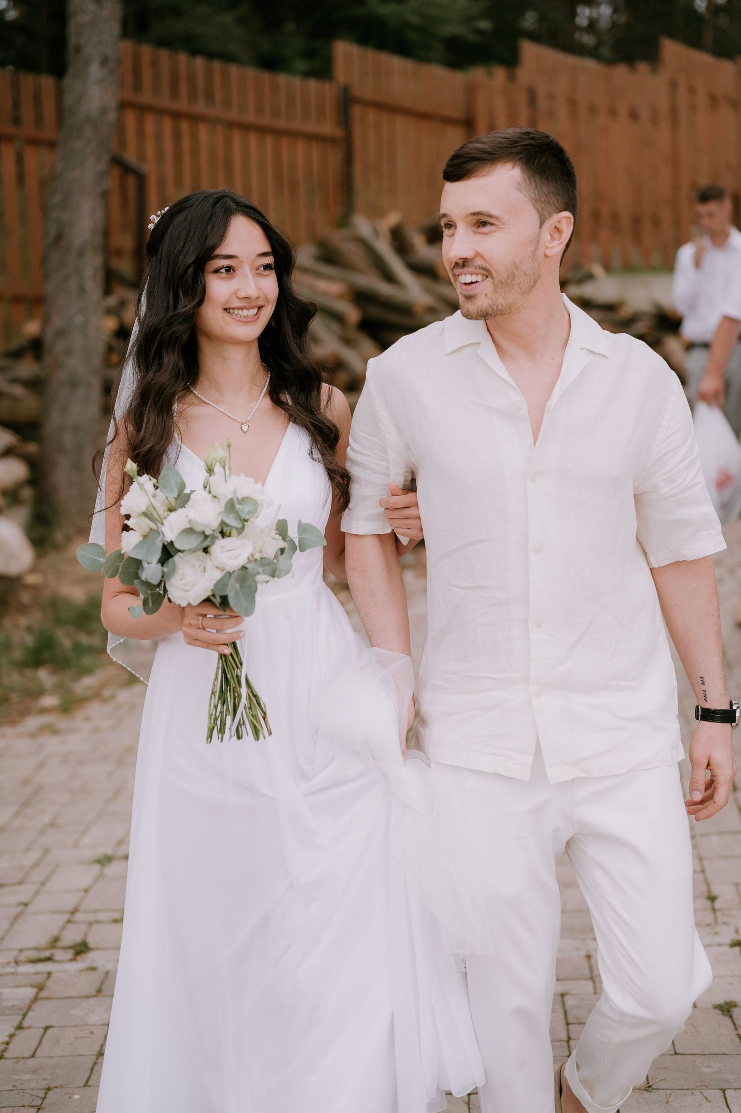 Couple in all-white attire walking together on stone pathway during wedding celebration.