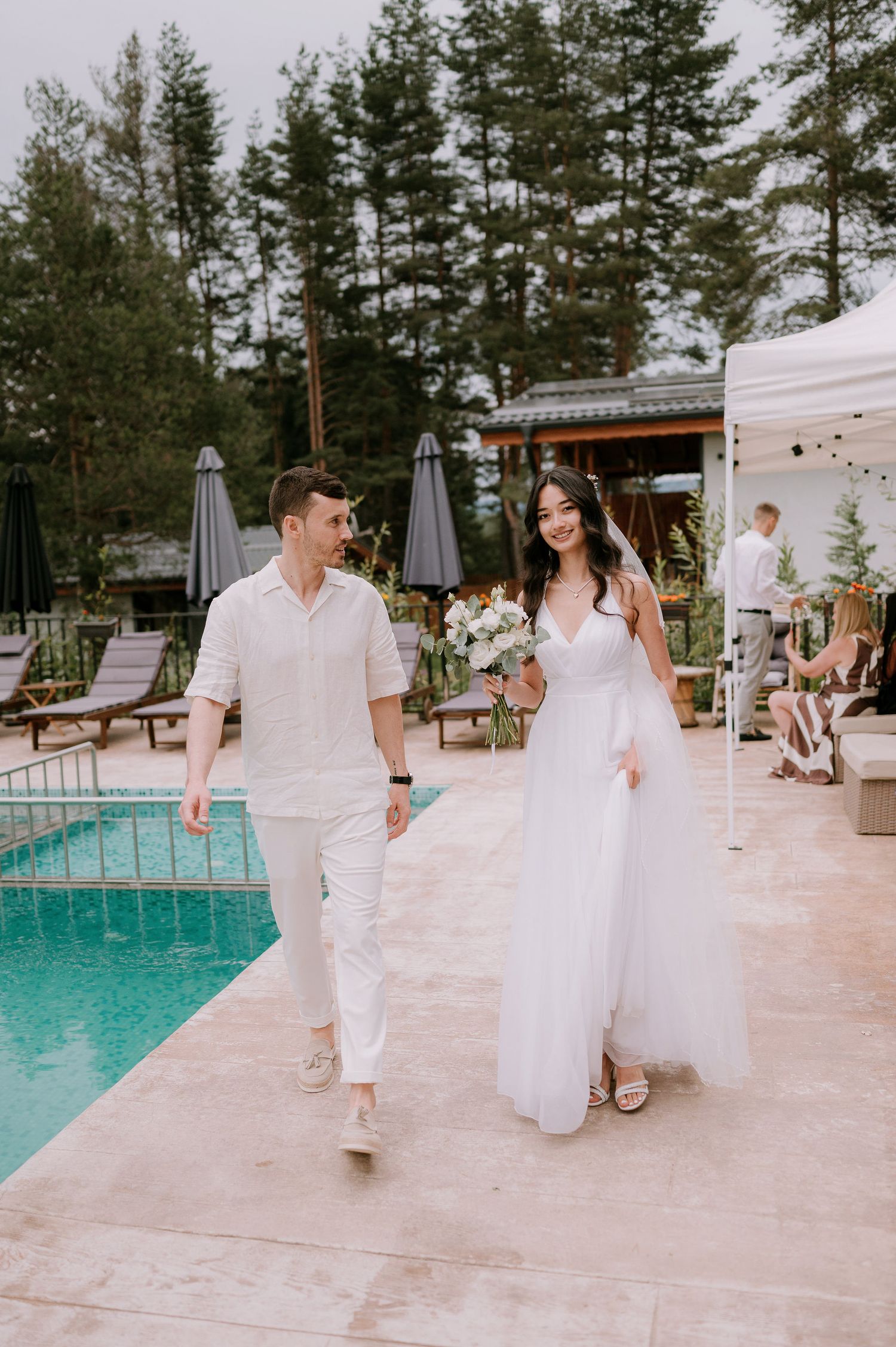 Wedding guests in white attire mingle on a stone patio beside a luxurious swimming pool.