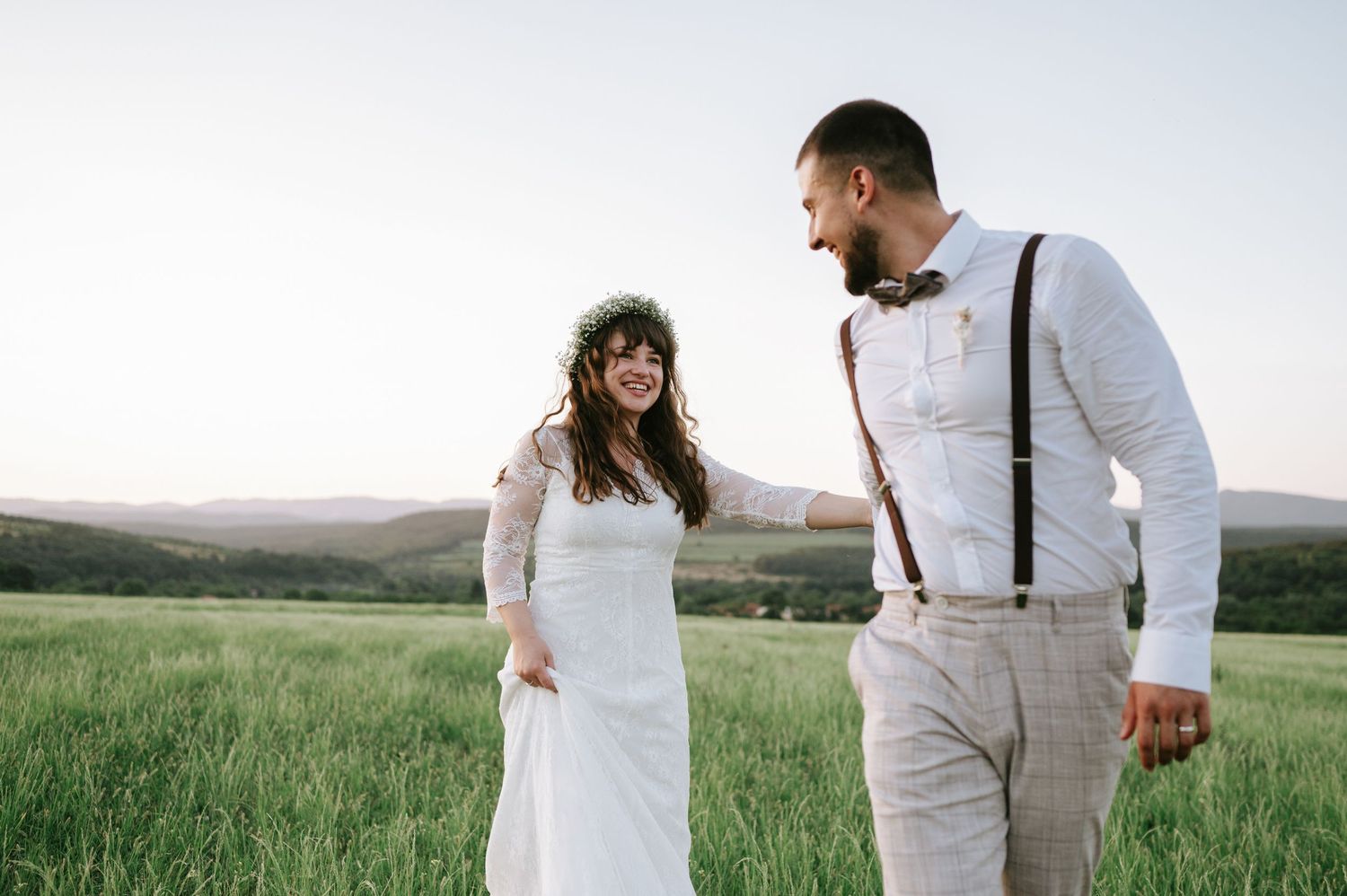 A couple in white outfits walks through a scenic green meadow with rolling hills in the background.