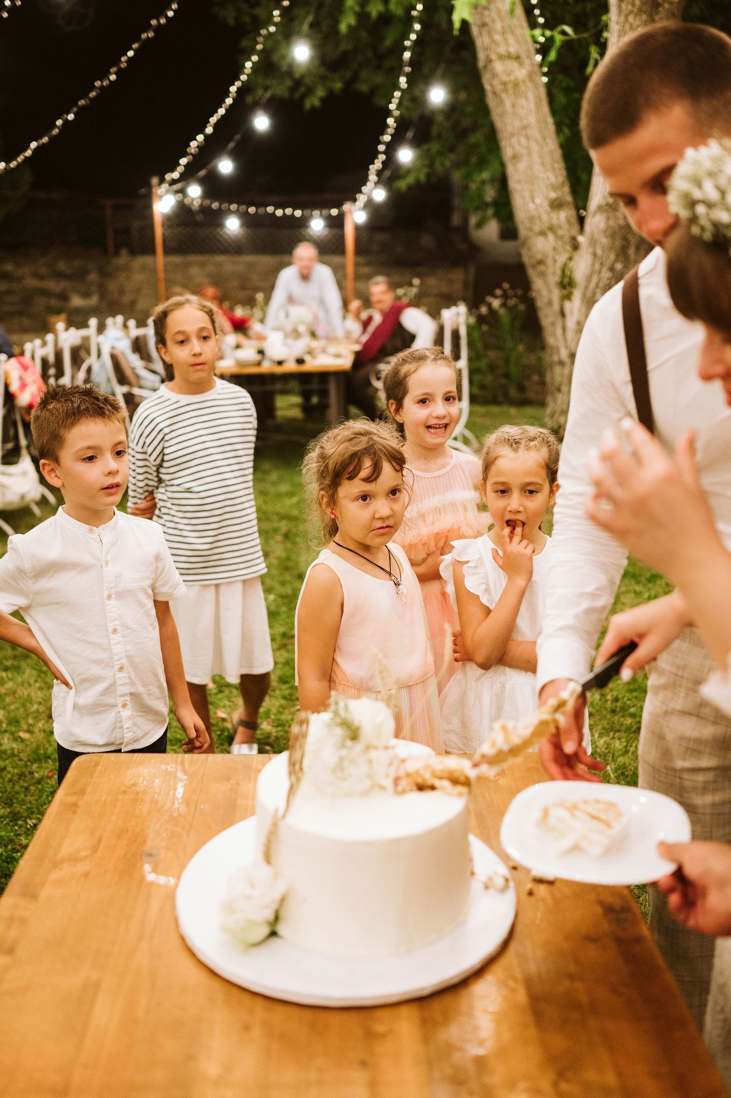 Children celebrate around a wedding cake at a rustic outdoor reception.