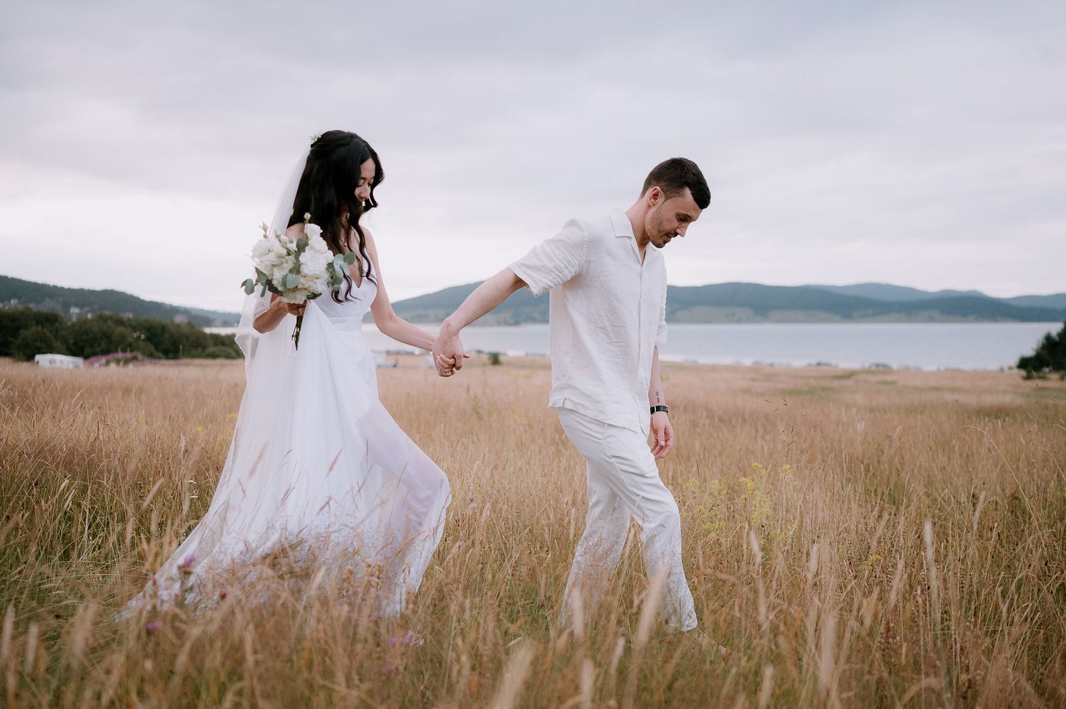 Couple in white outfits walking hand in hand through coastal meadow.