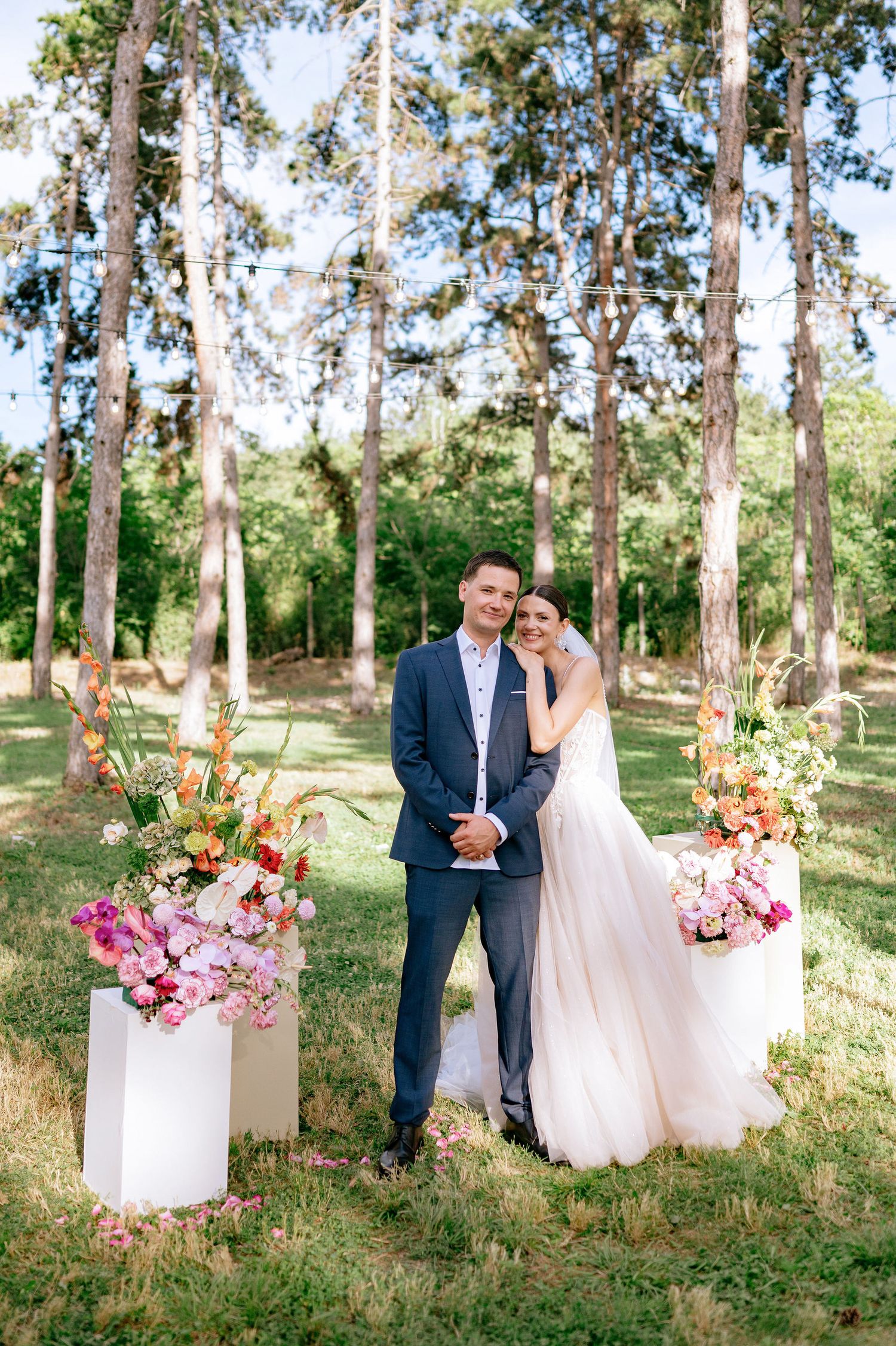 An elegant wedding couple poses between white floral arrangements in a sunlit forest setting.