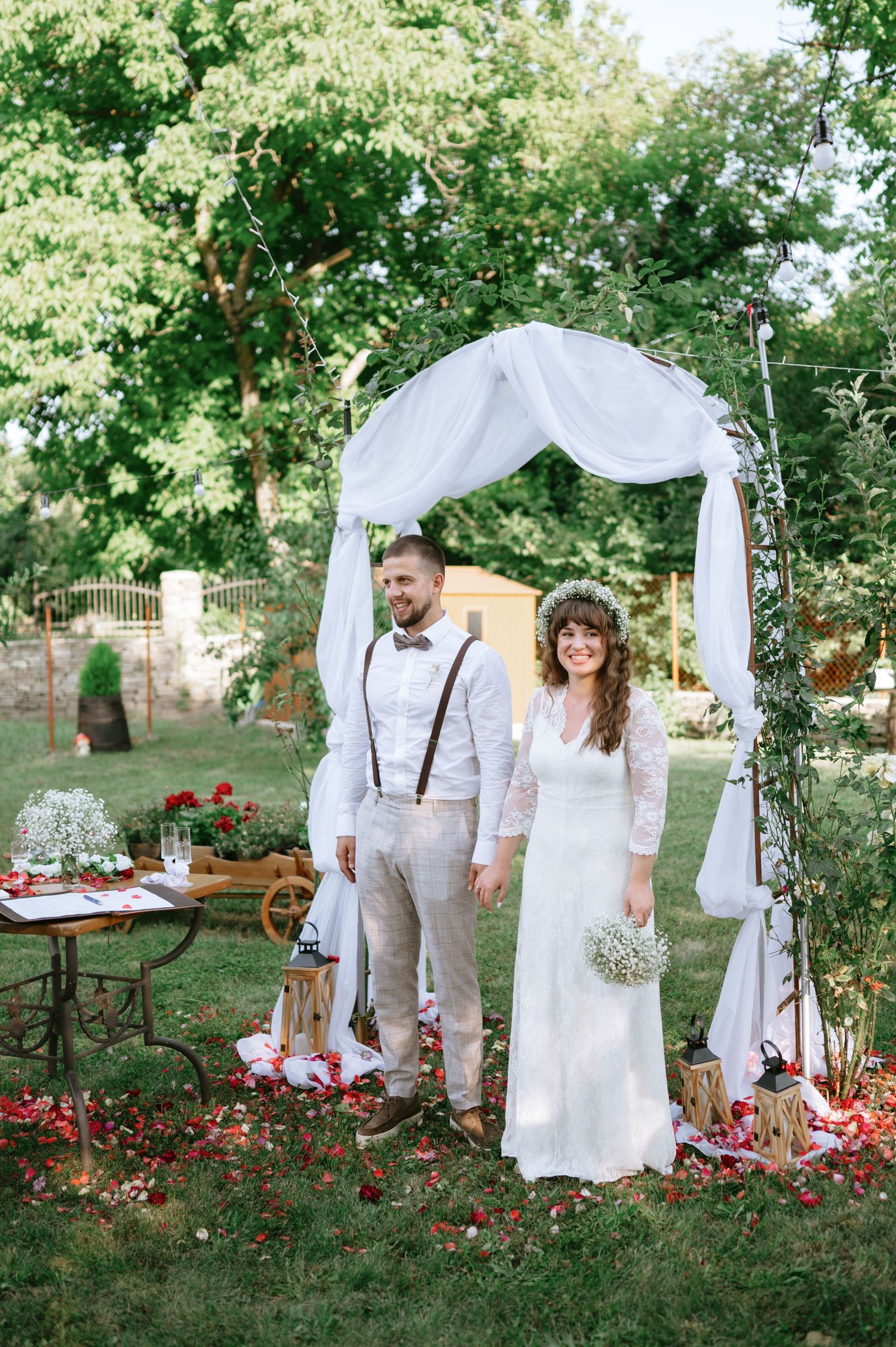 A garden wedding scene with a white fabric arch and red flower petals scattered on the grass.