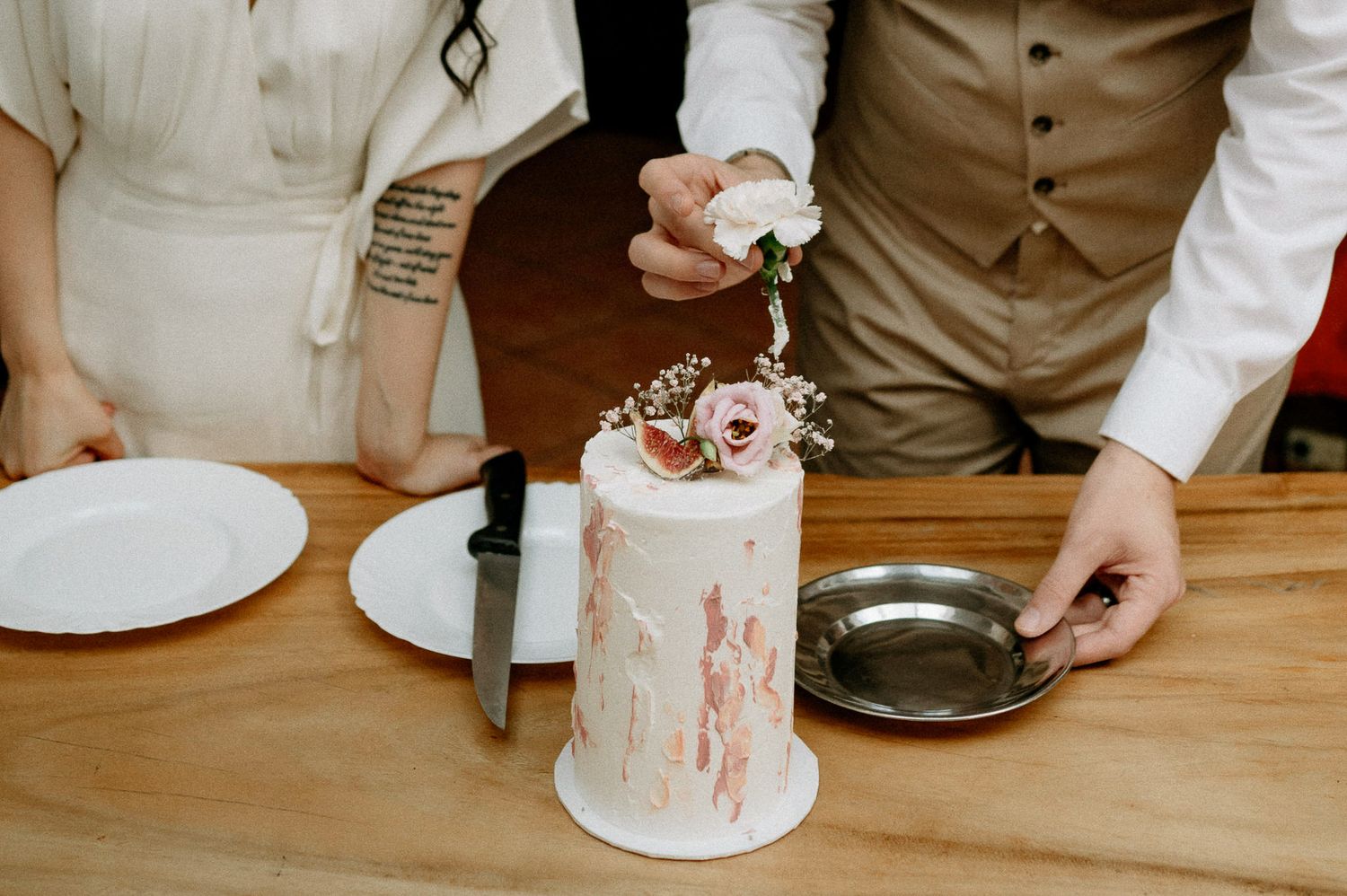 Wedding couple stands at wooden table with their small decorated cake.