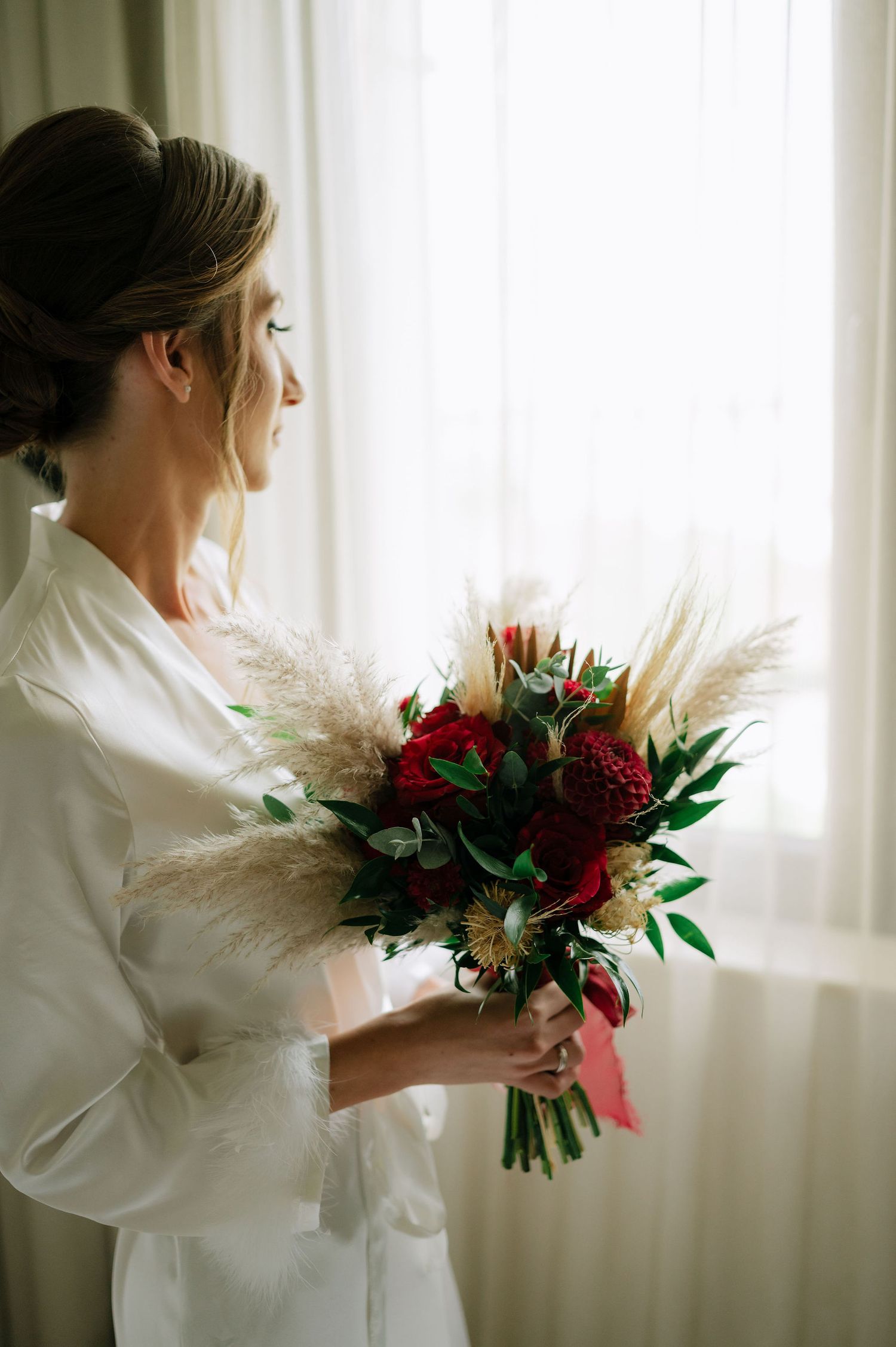Elegant figure in white robe holding a stunning red and cream wedding bouquet by window.