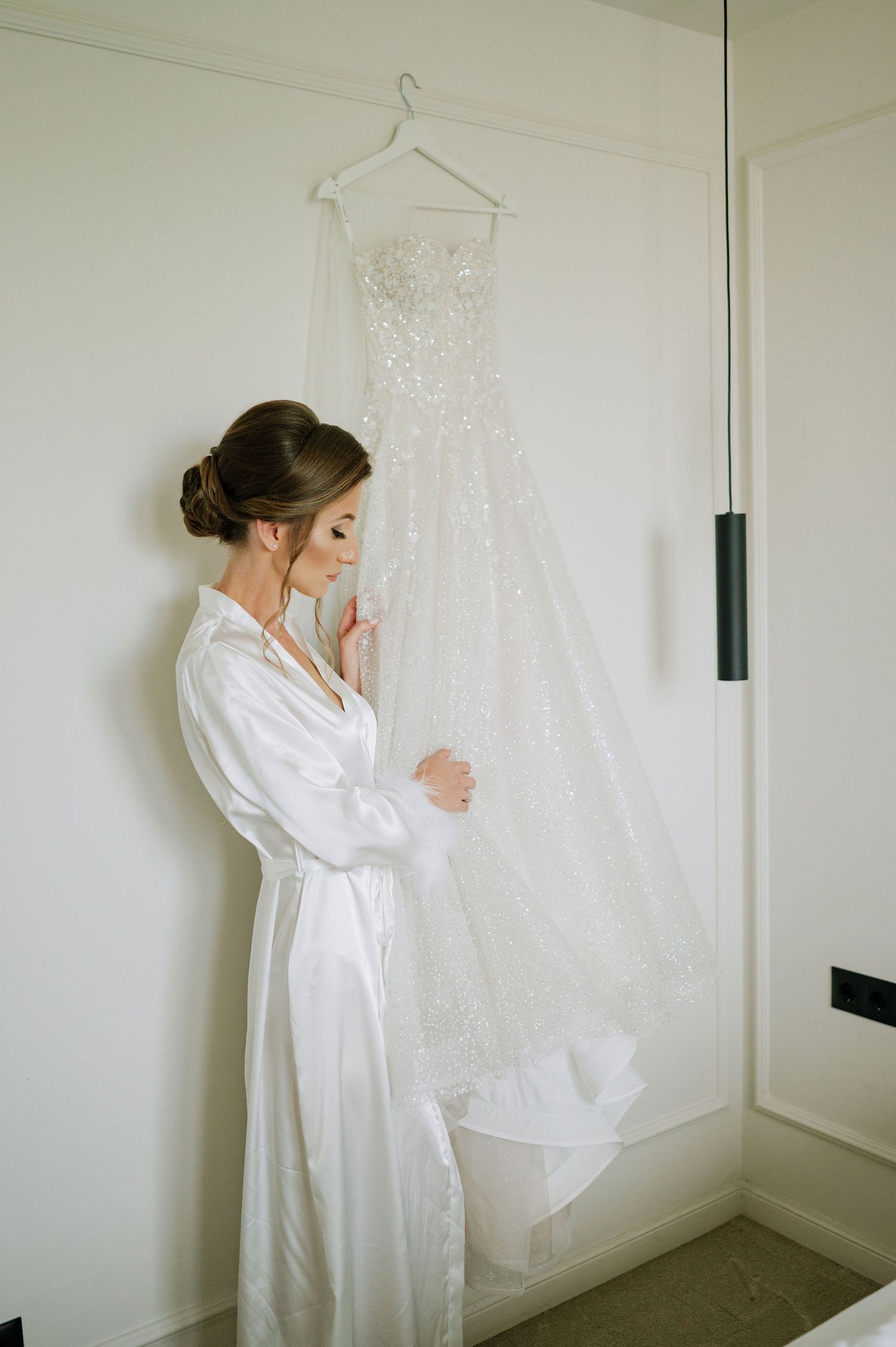 Wedding dress displayed on wall as figure in white robe admires it.