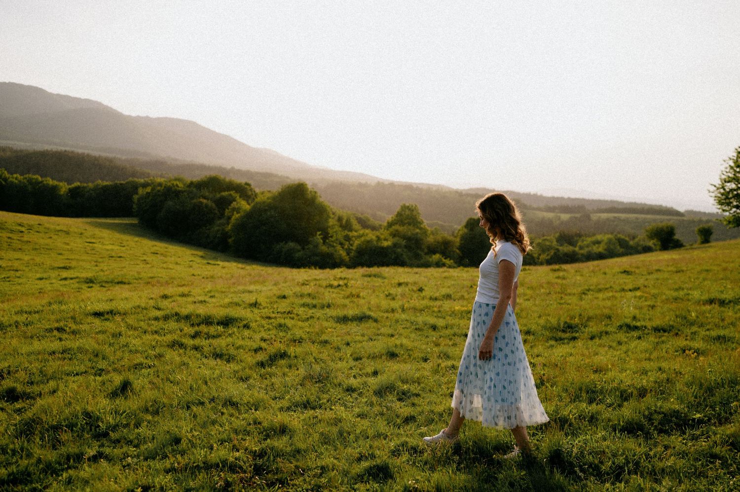 A person dances freely in a polka dot dress on a grassy hillside during sunset.