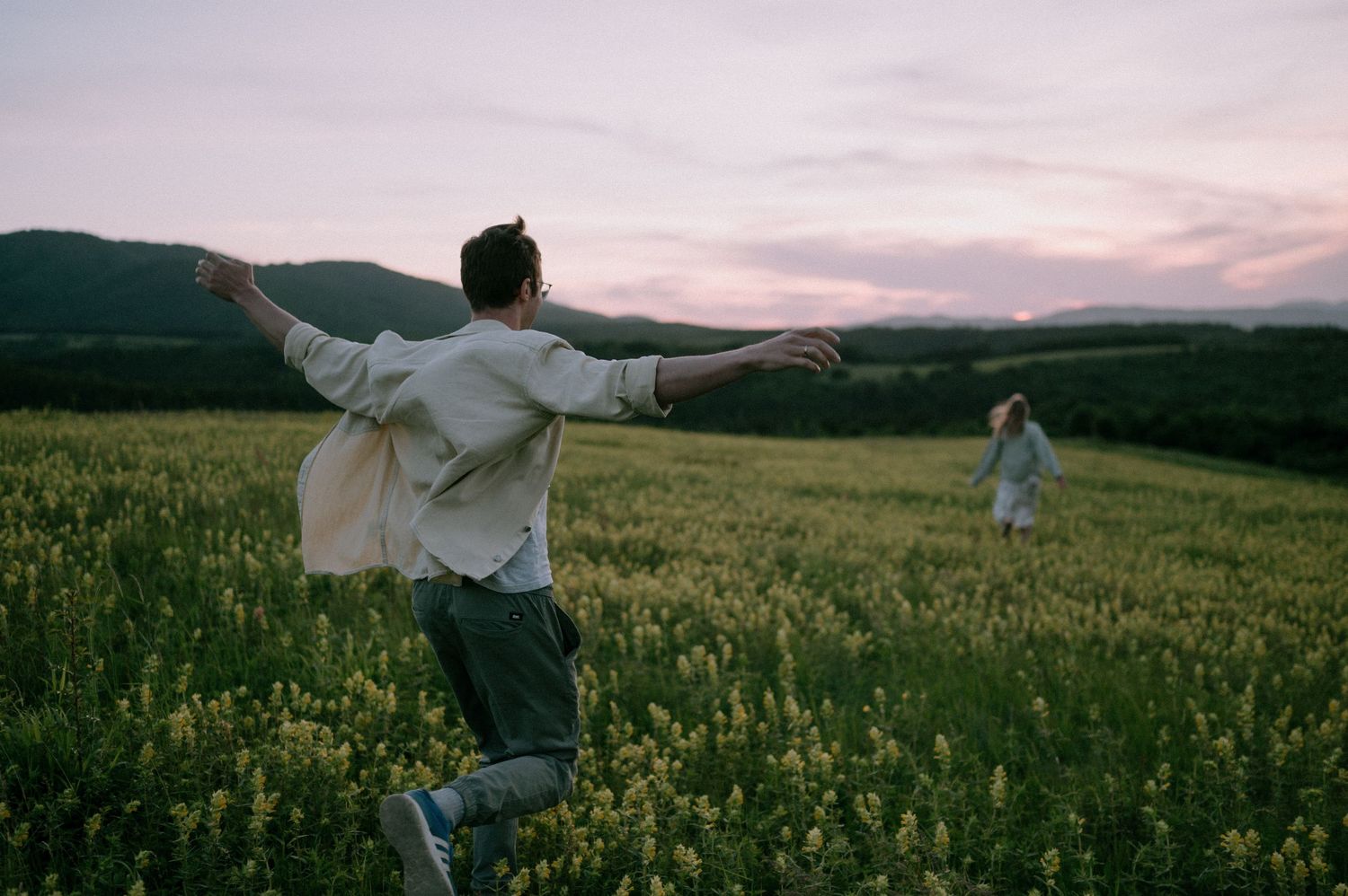 Two people dance through a vast field of yellow wildflowers with rolling hills in the distance.