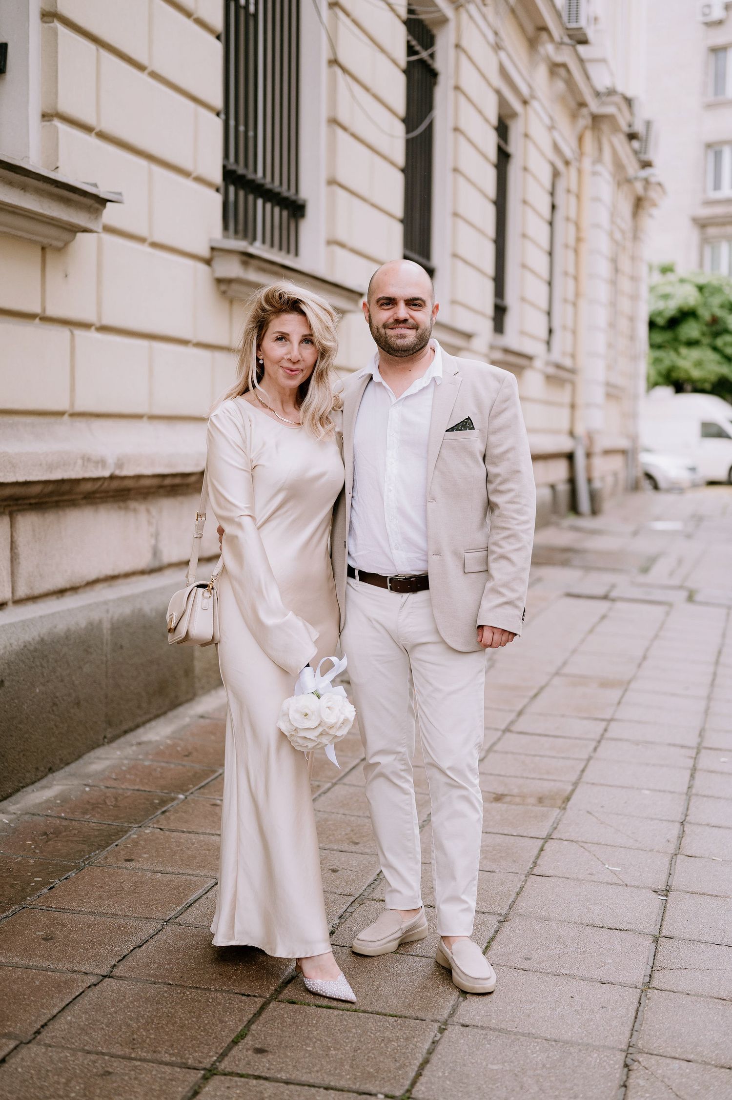 Bride and groom in all-white outfits share intimate moment by historic building.