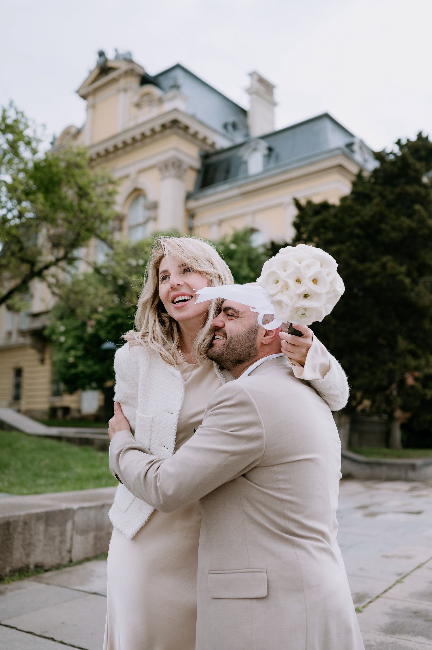 A couple in cream outfits share playful moments during their photoshoot near a historic building.