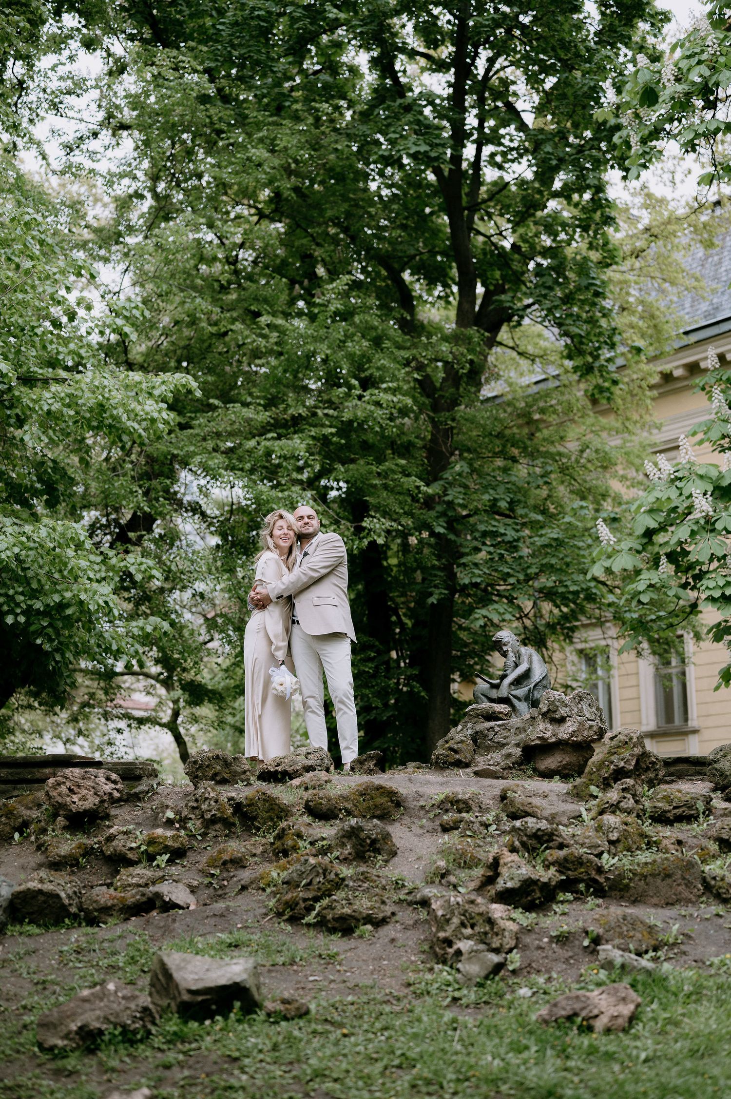 A couple in light-colored outfits embrace on rocky terrain near a historic building.