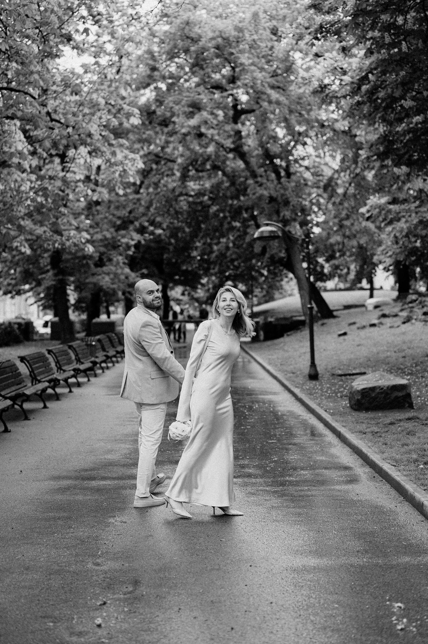 A romantic couple strolls down a park pathway surrounded by lush greenery and benches.