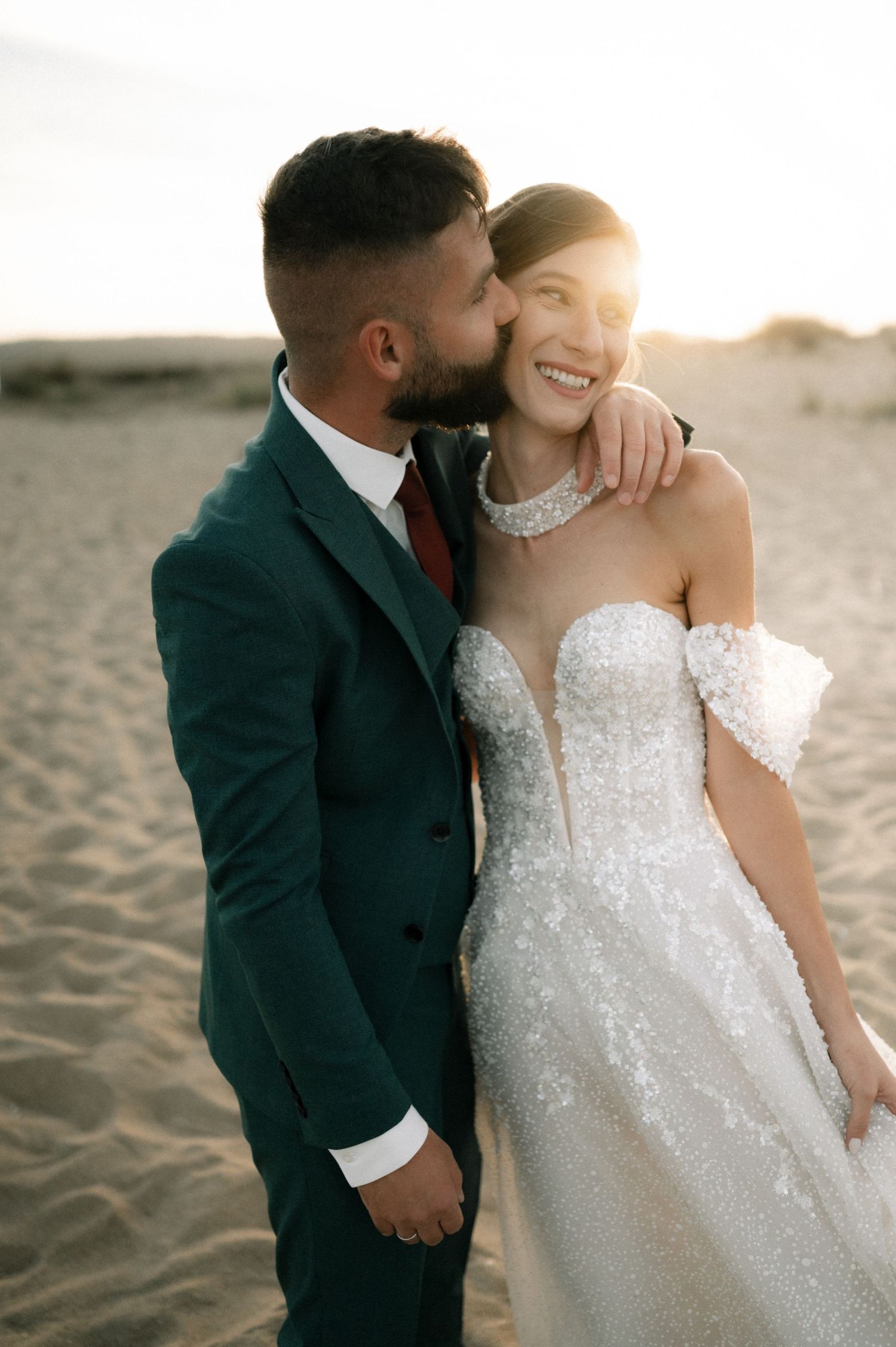 Bride and groom share a romantic sunset kiss on sandy dunes, she wears a strapless lace gown, he wears a dark green suit.
