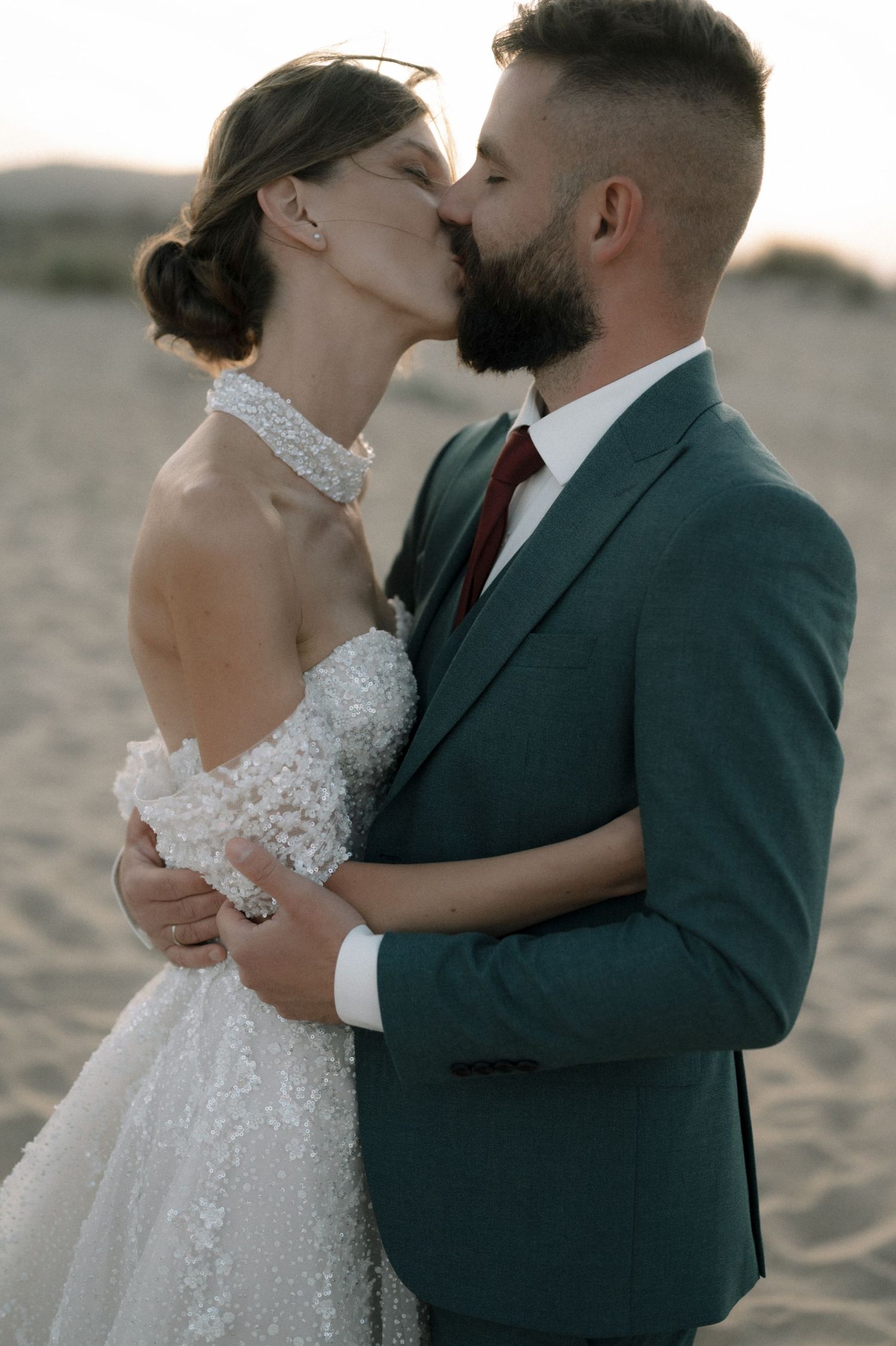 A bride and groom share a romantic kiss on a sandy beach, she in a lace gown, he in a teal suit with burgundy tie.