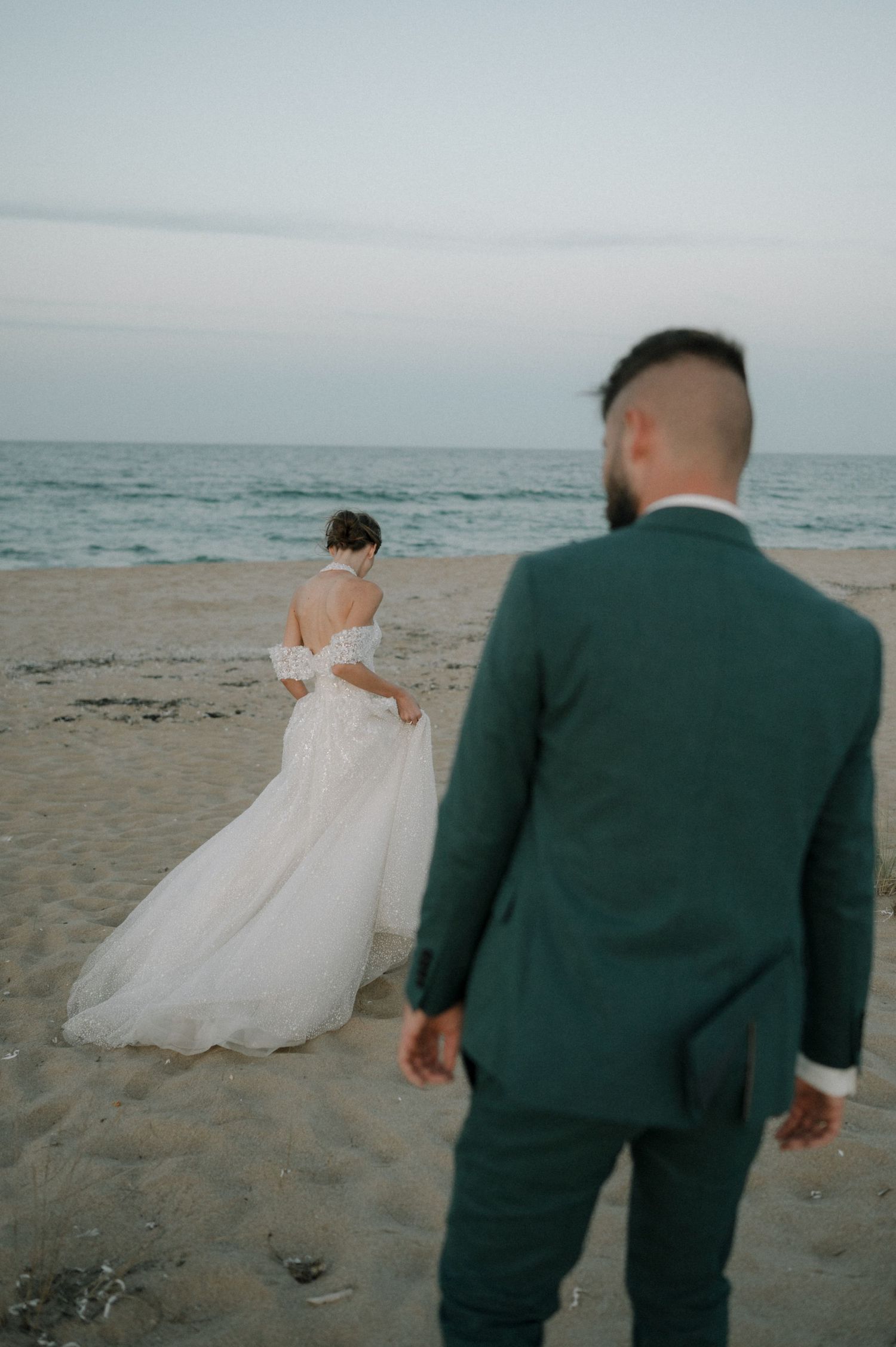 Bride in white off-shoulder gown and groom in green suit stand on a sandy beach at dusk near the ocean.