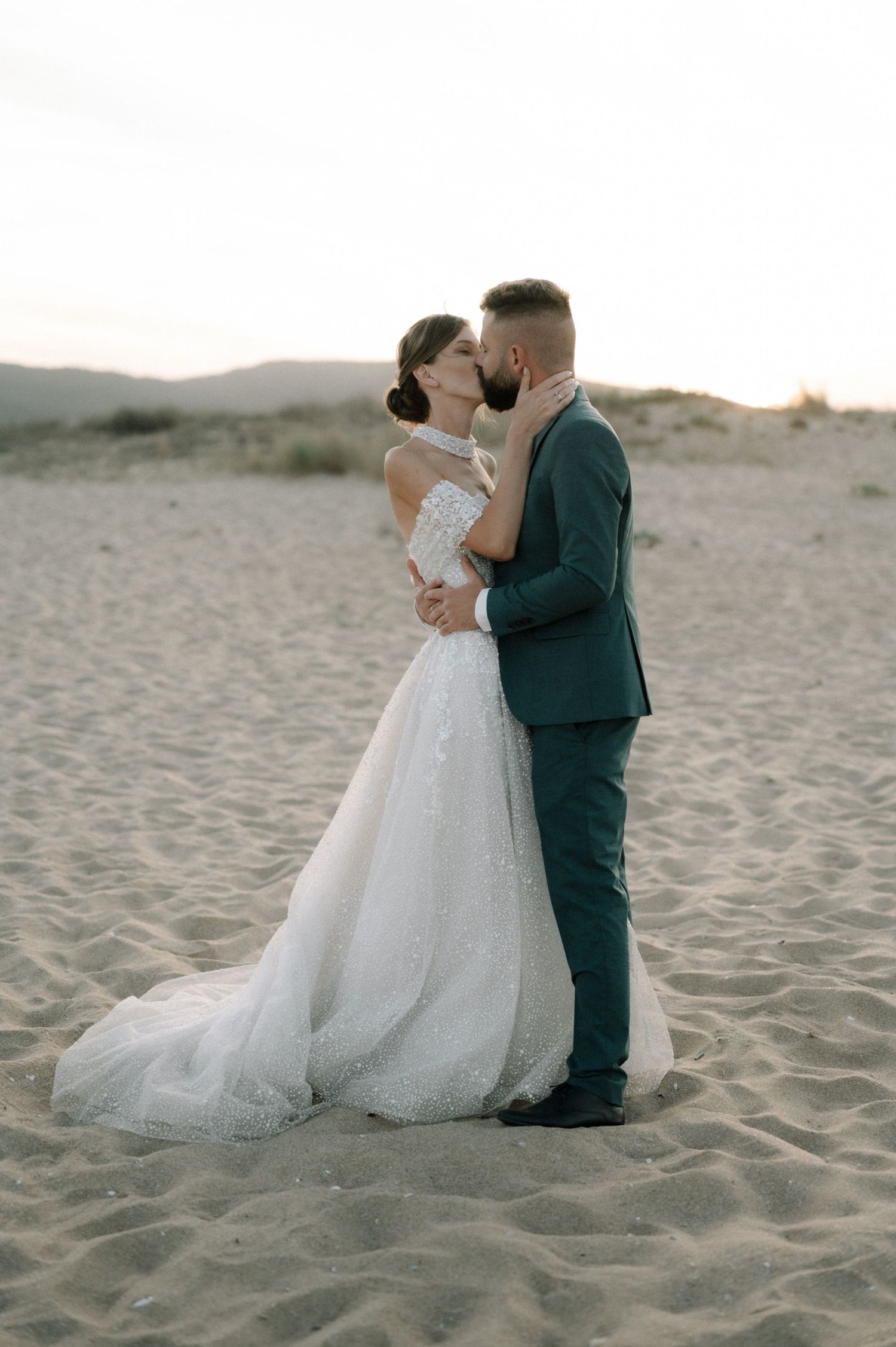 A bride and groom share a romantic kiss on a sandy beach at sunset, she in a white gown, he in a dark suit.