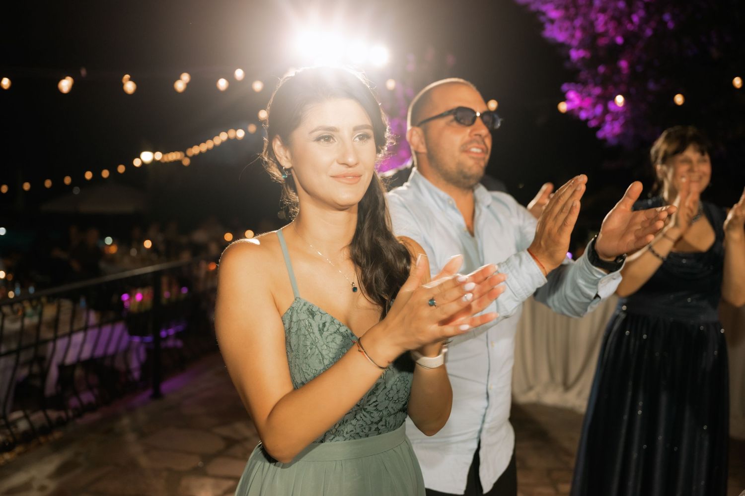 Wedding reception guests celebrate under string lights and purple uplighting while dancing at an outdoor venue.