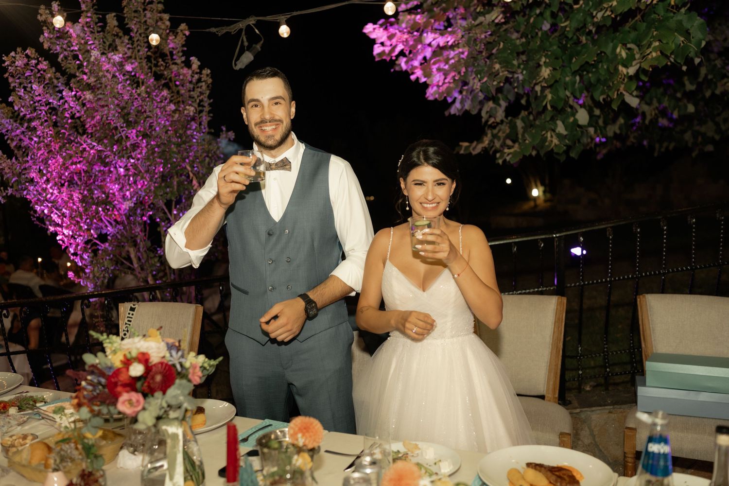Wedding reception dinner outdoors with purple bougainvillea flowers and string lights creating a romantic ambiance.