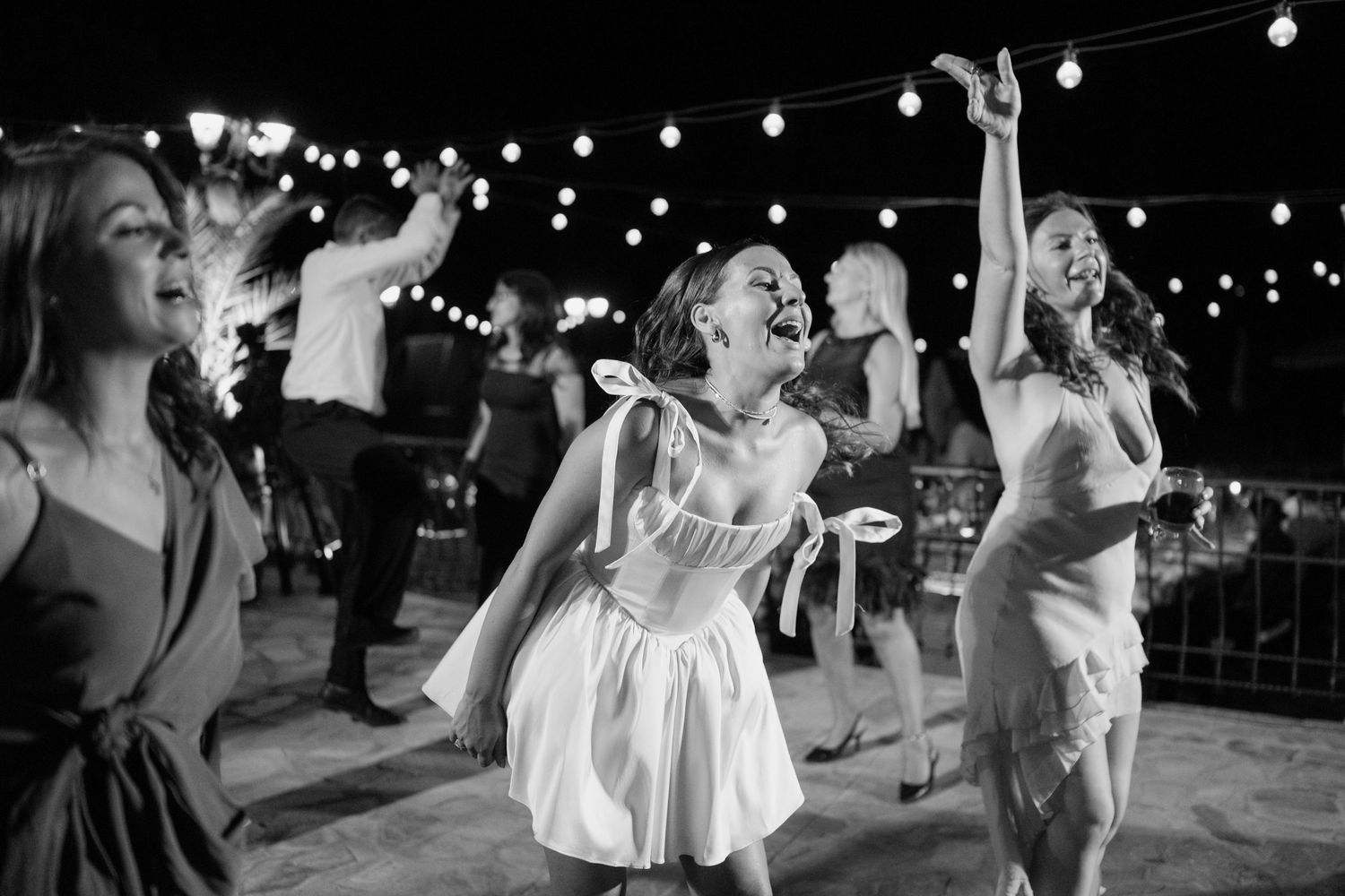 Wedding guests dance joyfully under string lights at an outdoor evening reception celebration.