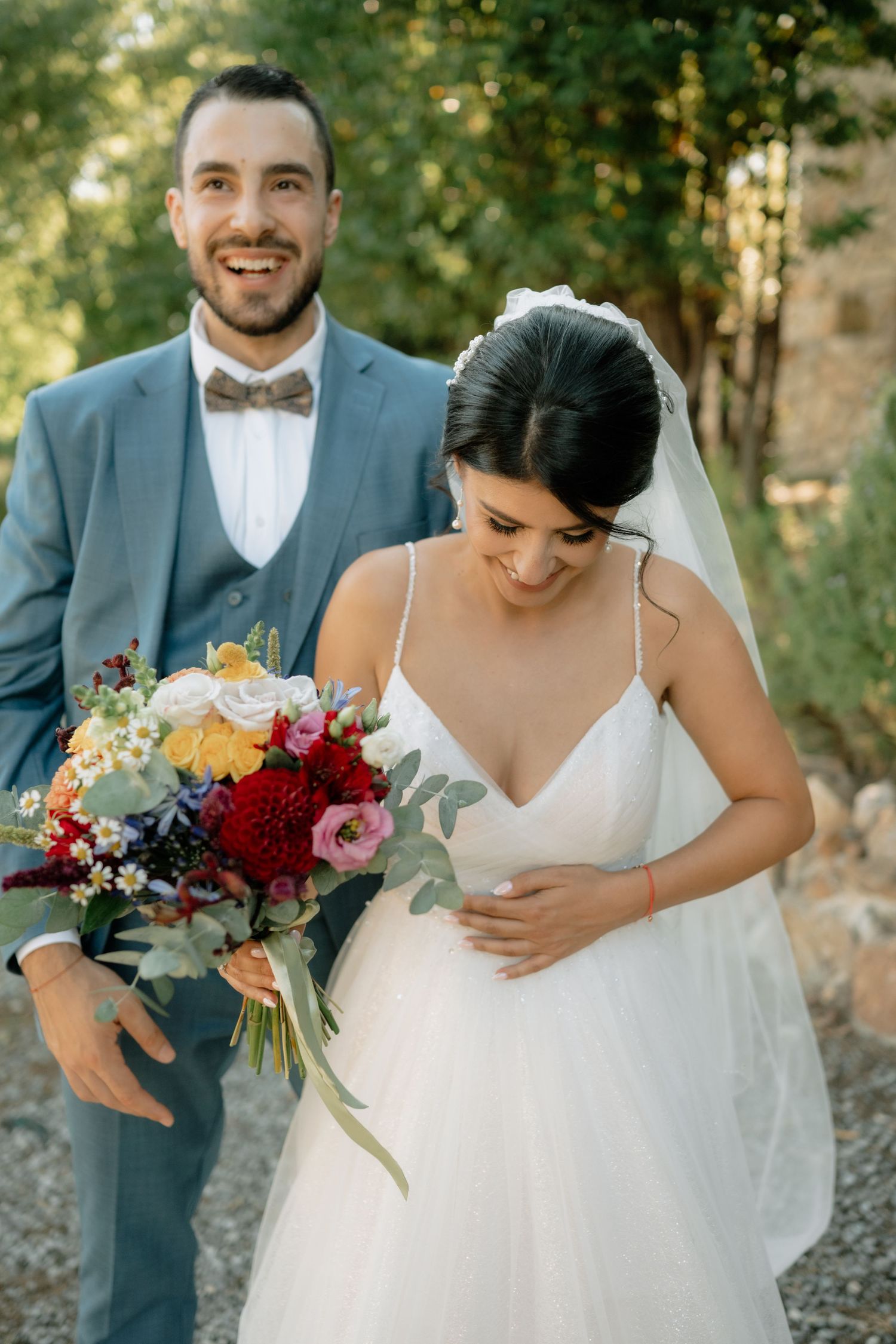 Wedding couple and wedding party members in gray suits hold vibrant red and pink floral bouquets in outdoor setting.
