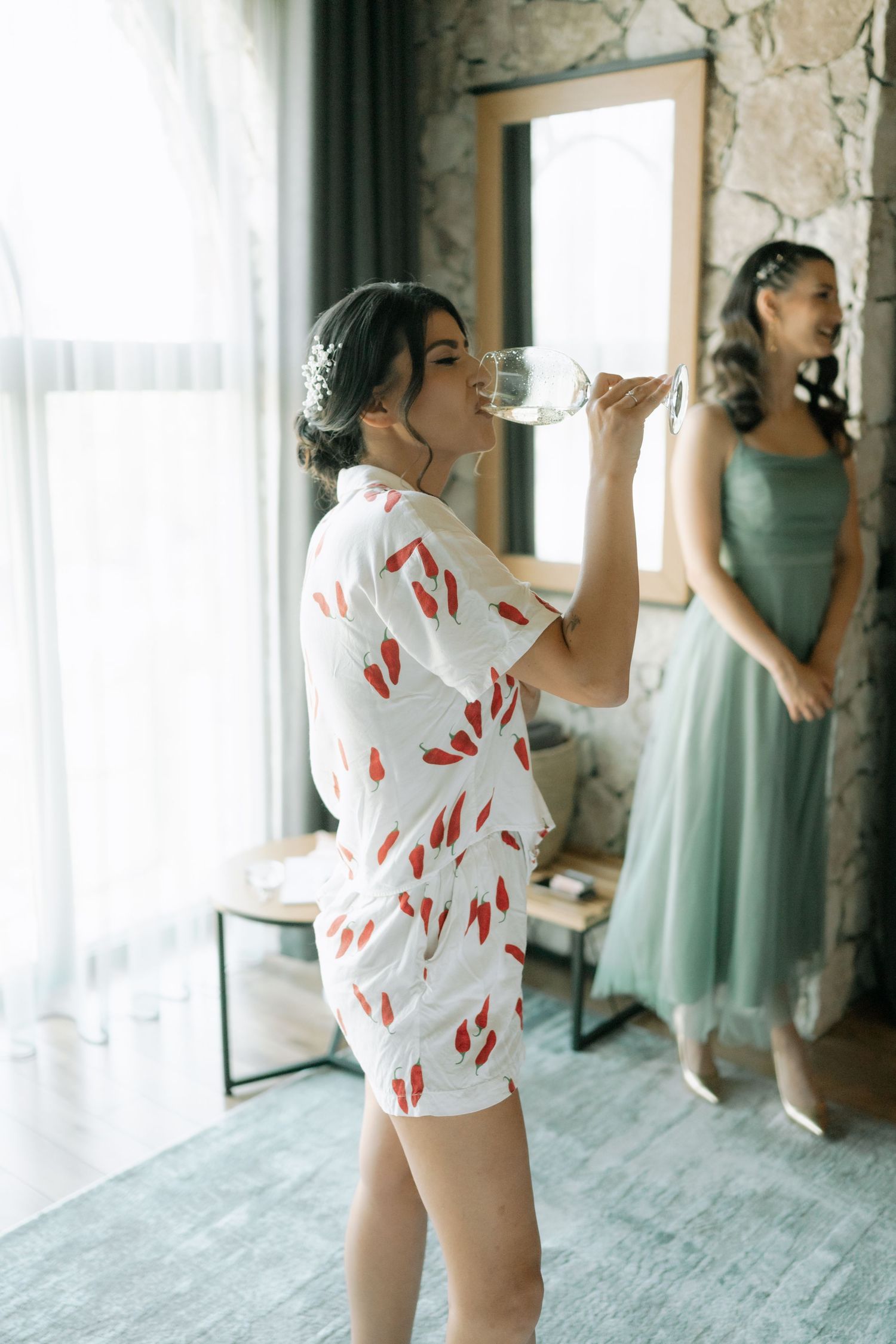 Two women getting ready in a room, one wearing a floral romper drinking from a glass, another in a mint green dress.