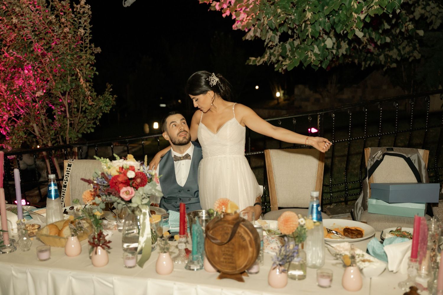 A wedding reception table decorated with floral centerpieces and place settings glows warmly in evening light.