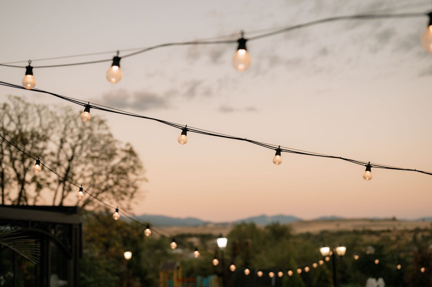 String lights hang across a dusky sky with silhouetted trees and mountains in the background at sunset.