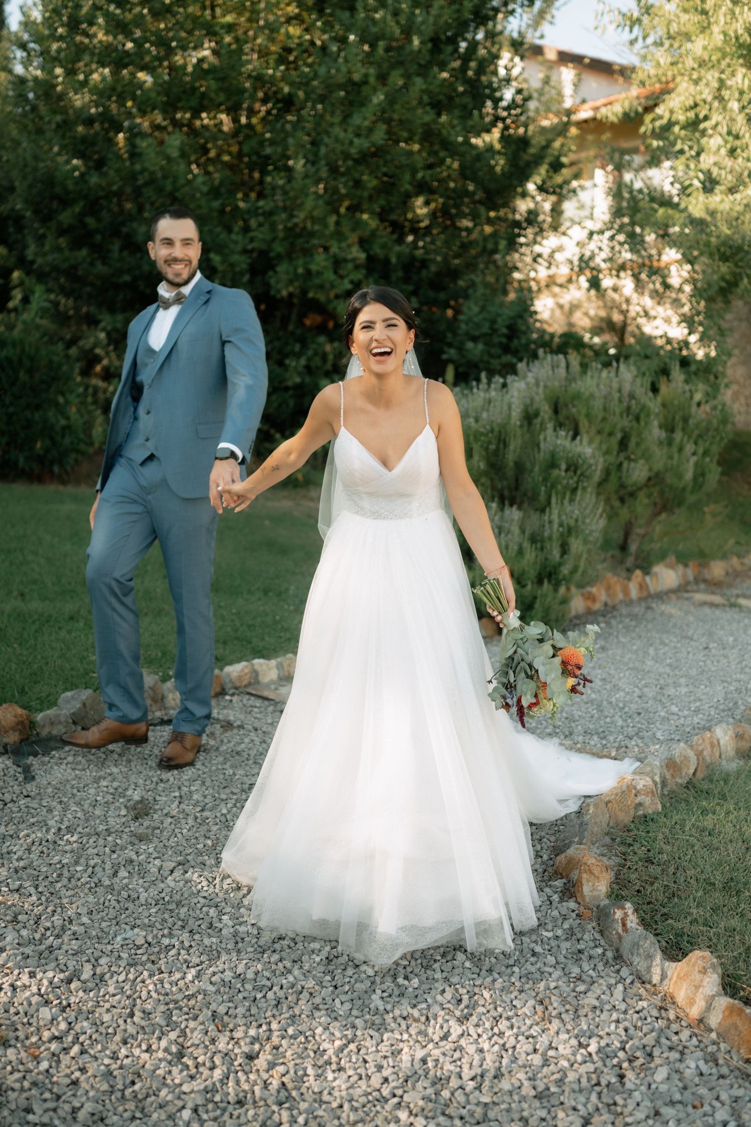 Wedding couple poses playfully on a gravel path surrounded by lush greenery in a romantic outdoor setting.