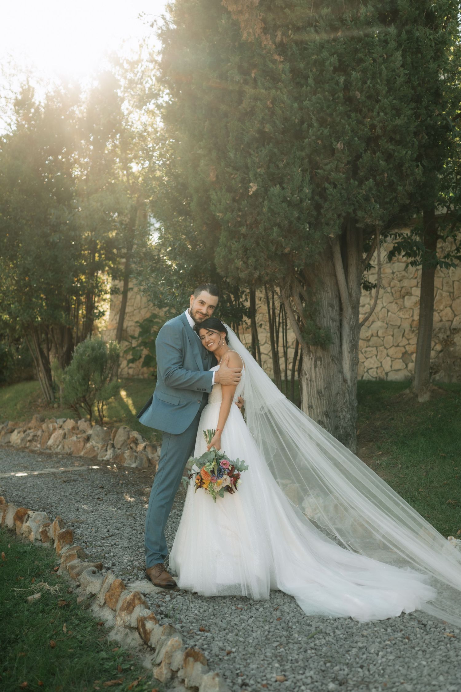 A couple in wedding attire shares an intimate moment on a gravel path with sunlit trees in the background.