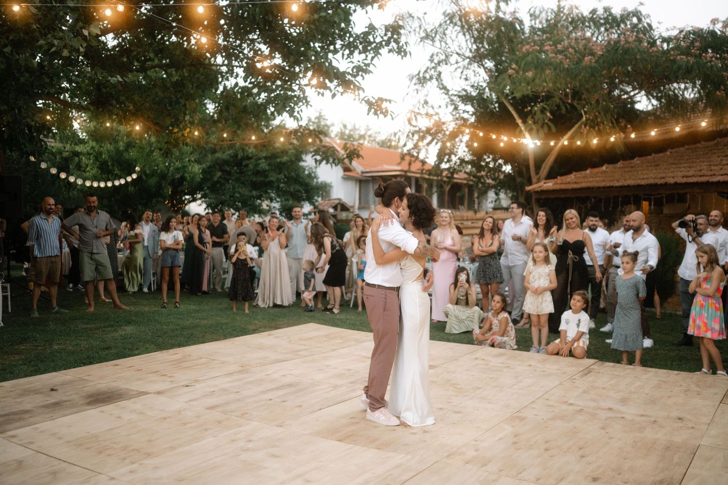 Wedding guests dance and celebrate under warm string lights at an outdoor evening reception.