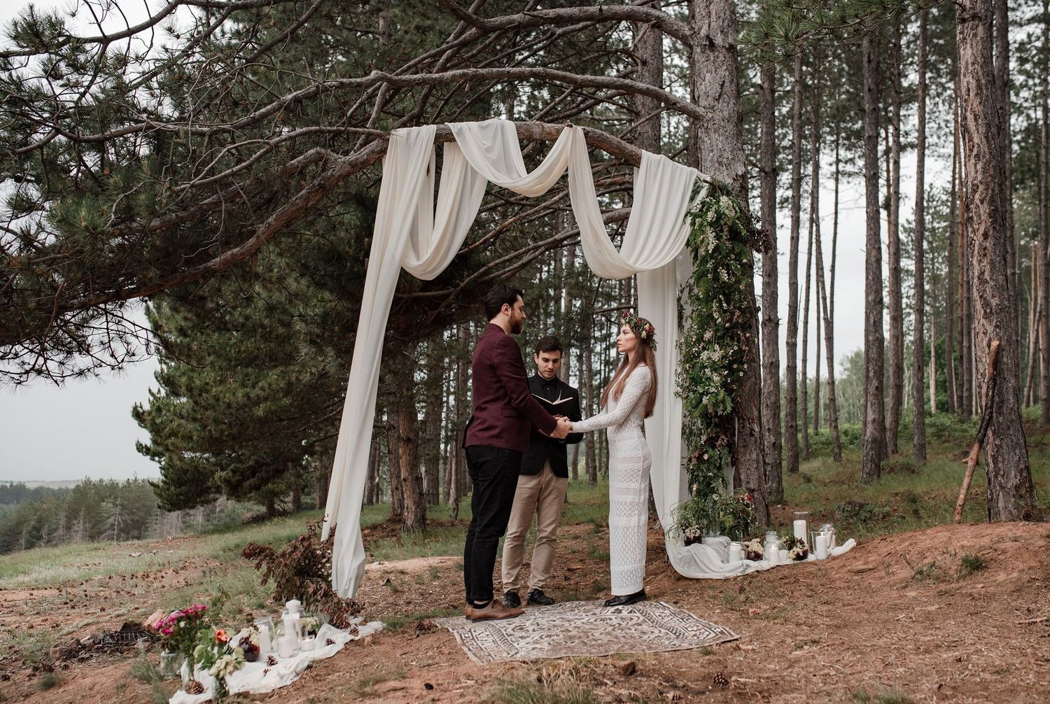 A winter wedding ceremony takes place in a snowy forest with evergreen trees and white fabric draped between trunks.