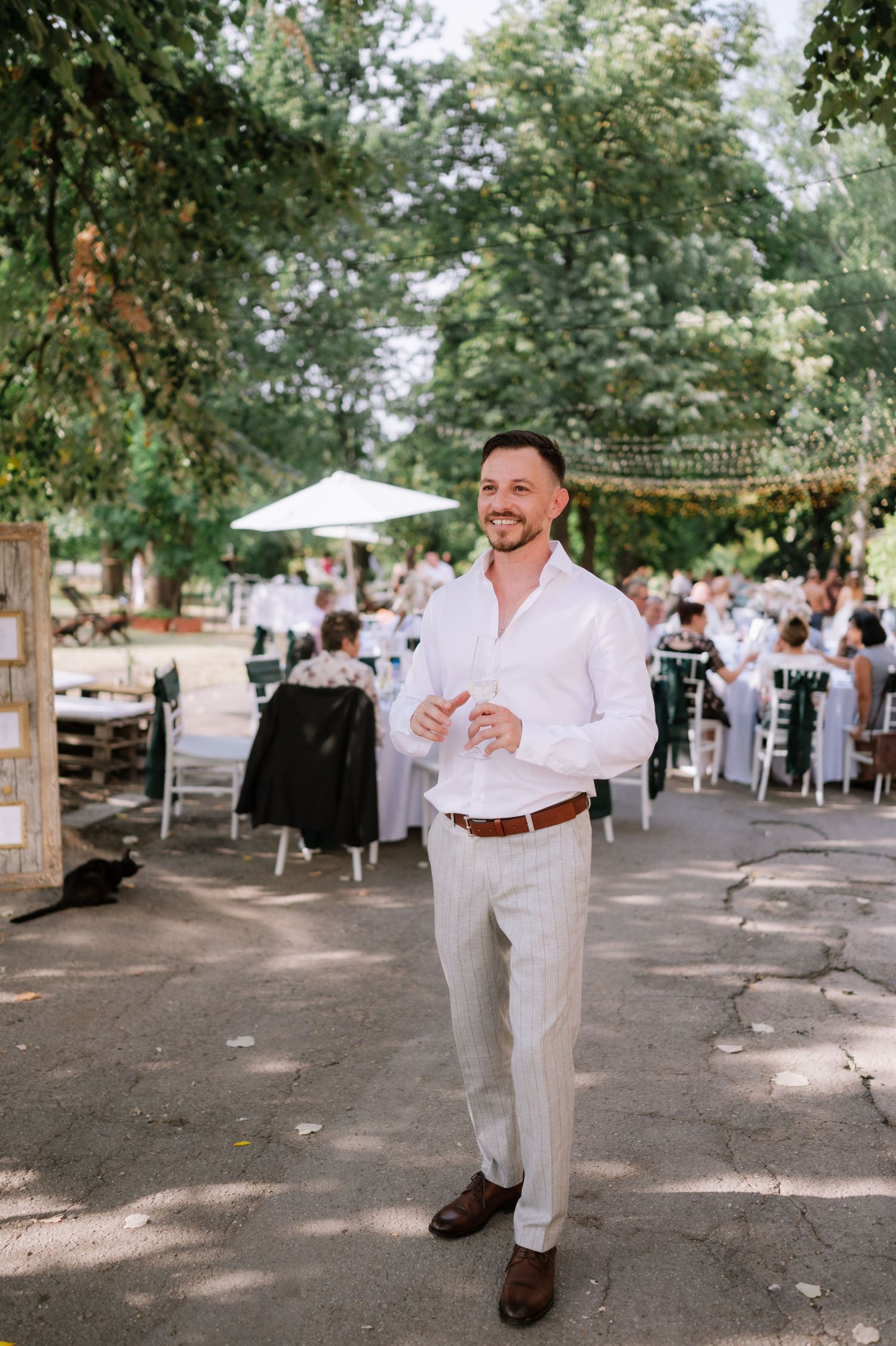 An outdoor wedding reception venue with string lights overhead and guests seated at tables under shaded trees.