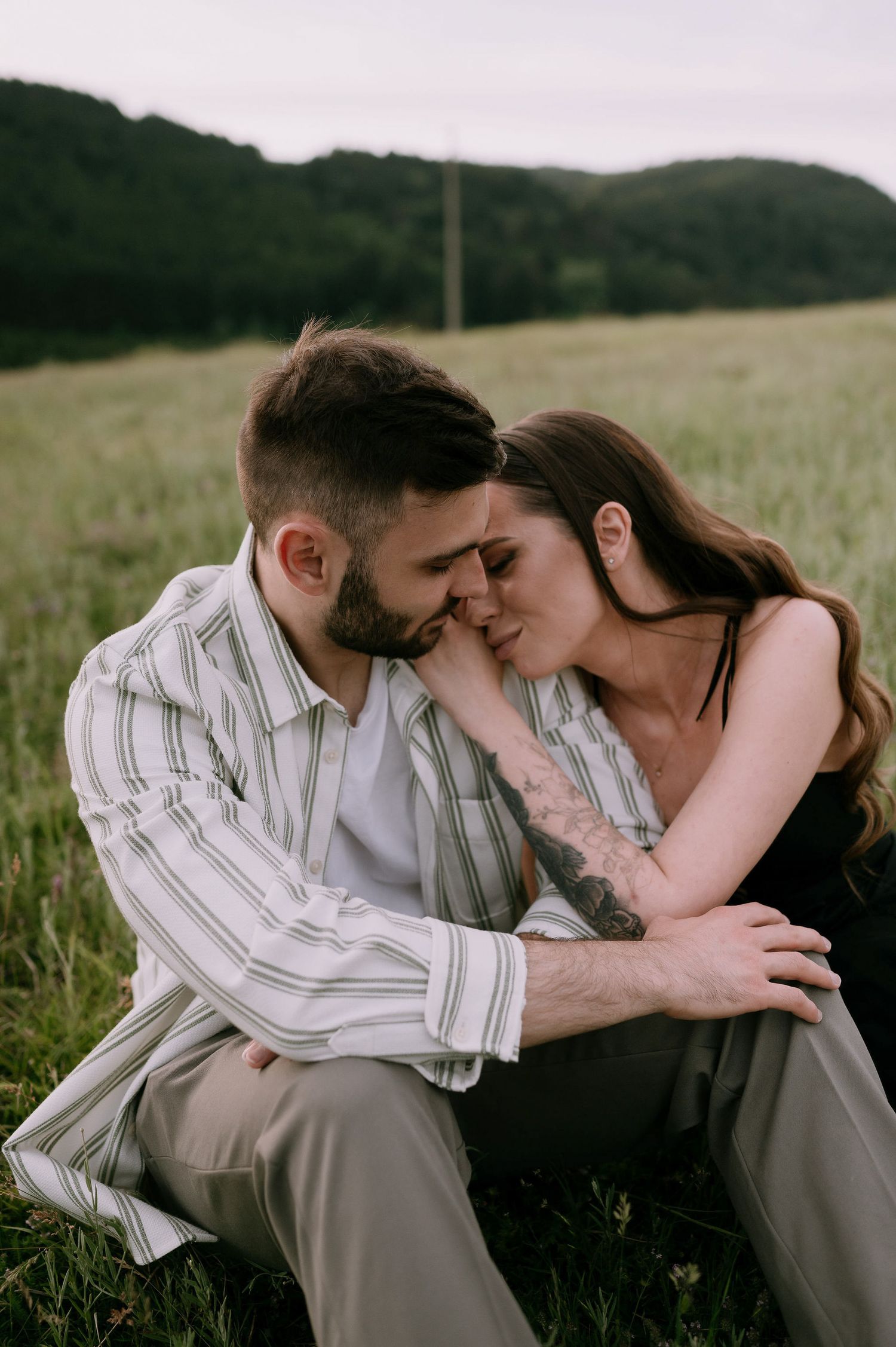 A romantic couple shares intimate moments in a grassy meadow with rolling hills in the background during golden hour.