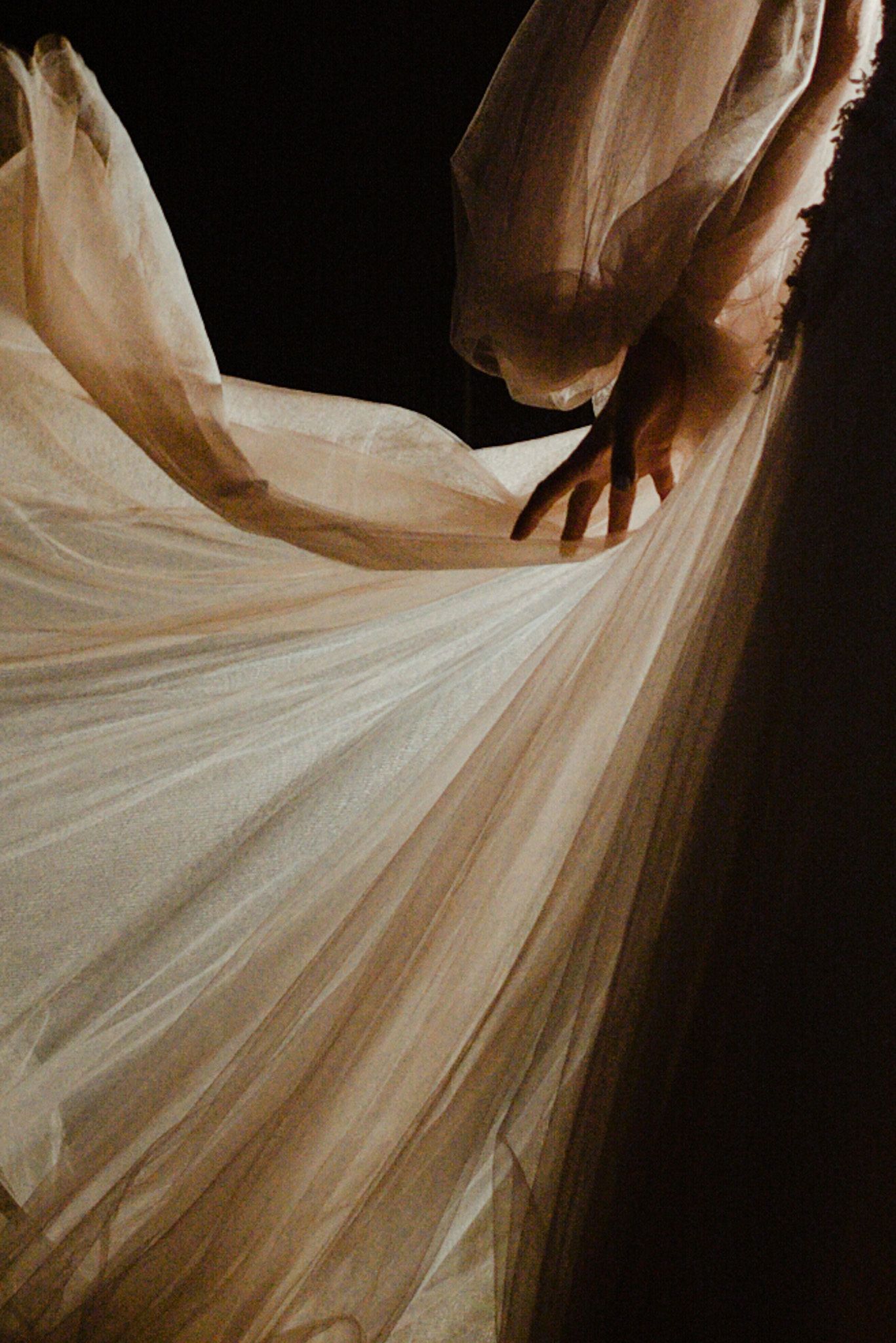 Elegant hands gracefully lift flowing white tulle fabric in an artistic dance movement against a dark background.