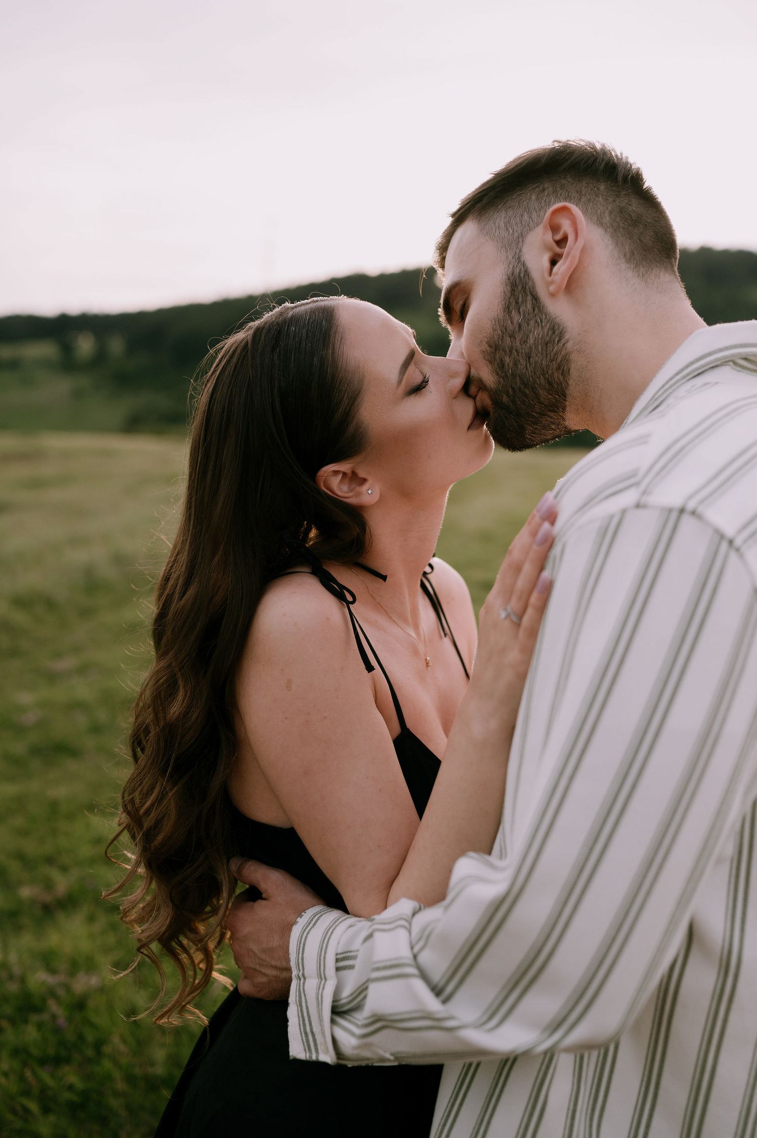 A couple shares an intimate kiss in a grassy field at sunset, with soft natural lighting creating a romantic atmosphere.