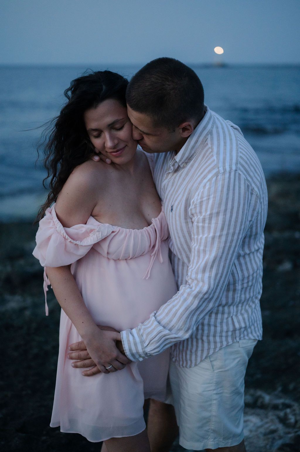 A couple in light colored clothing shares intimate moments by the ocean at dusk with a full moon rising in the background.