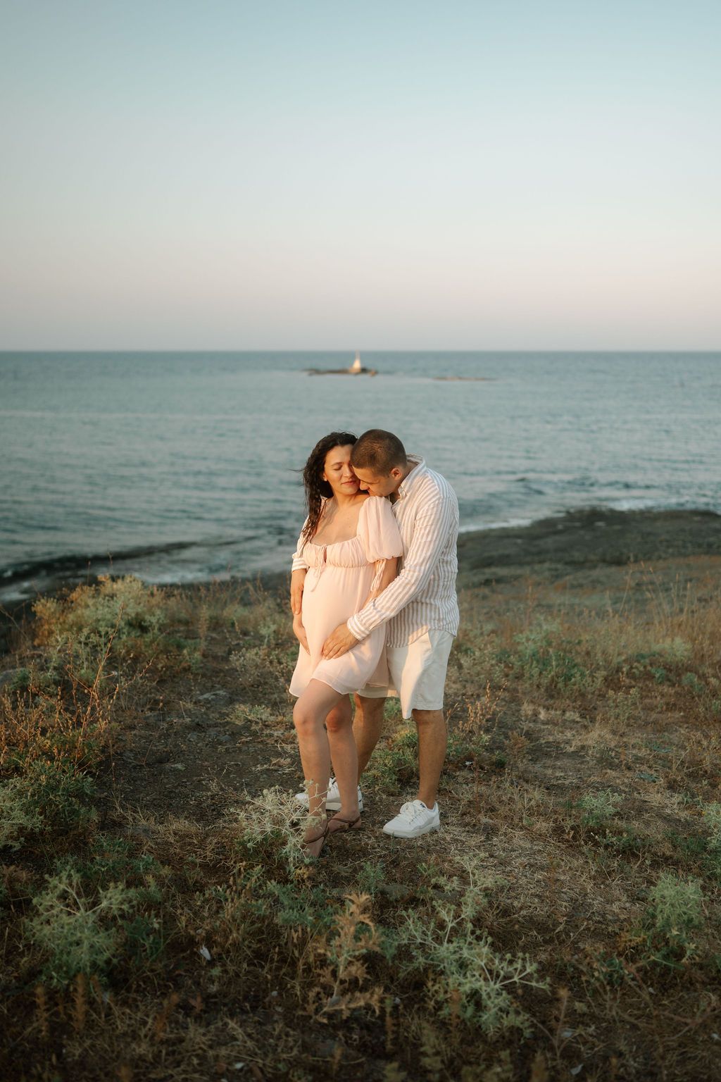 A romantic couple in white outfits embraces on a coastal cliff during golden hour as a sailboat passes in the distance.