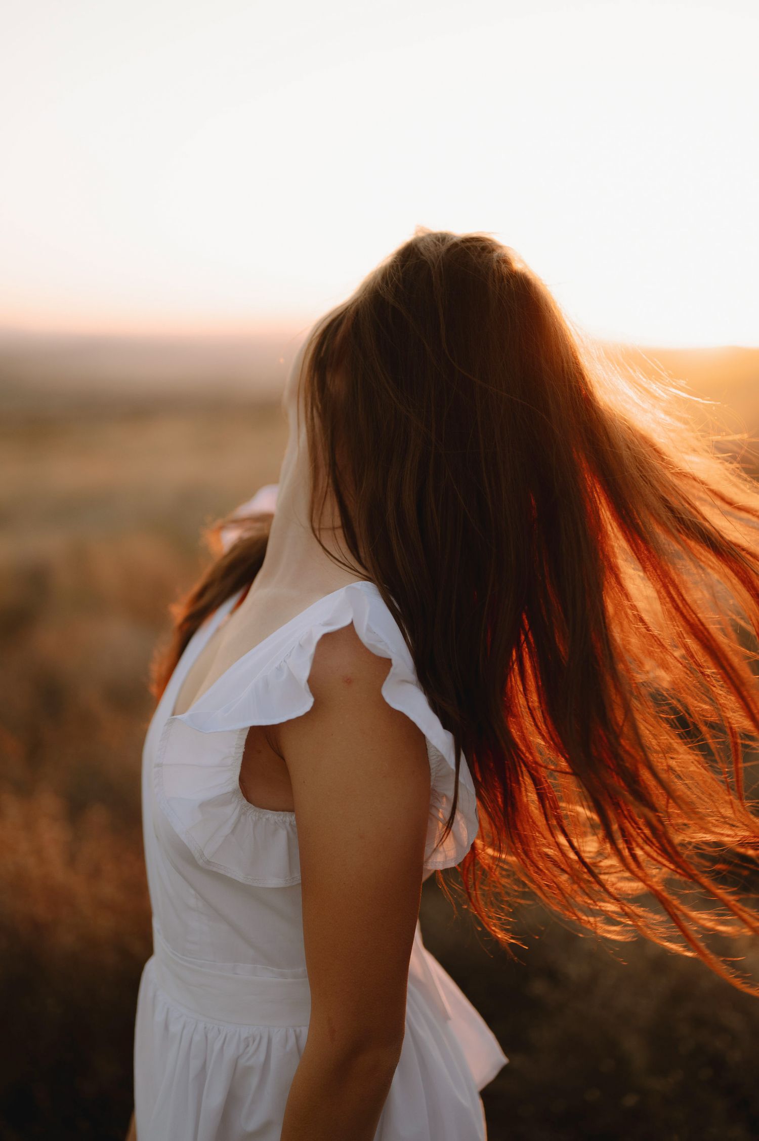 A person with flowing red hair in a white dress stands against a golden sunset landscape.