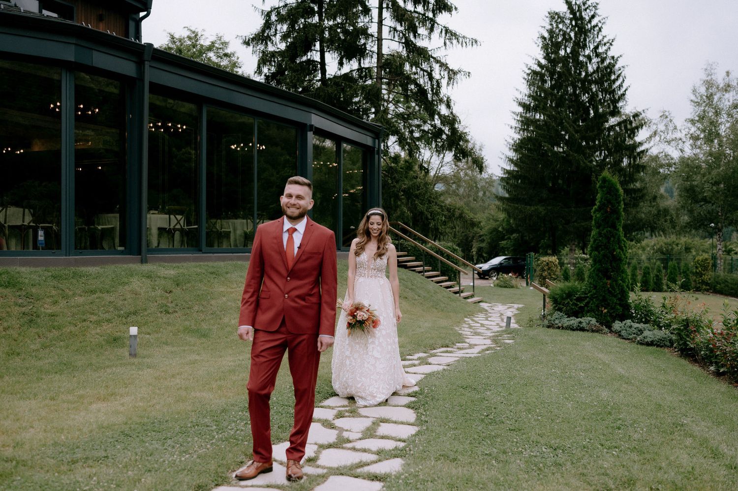 A couple in formal attire walks along a winding stone pathway through a lush green garden setting.