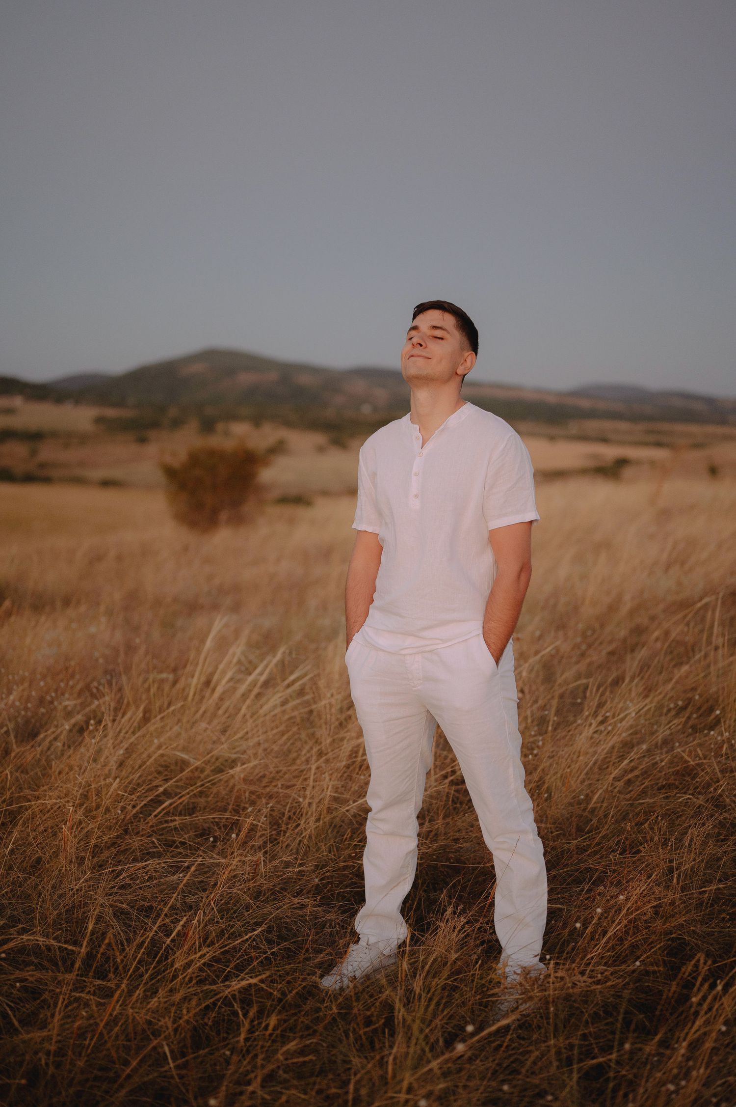 Figure in white clothing poses thoughtfully in golden grass field against mountain backdrop during sunset.