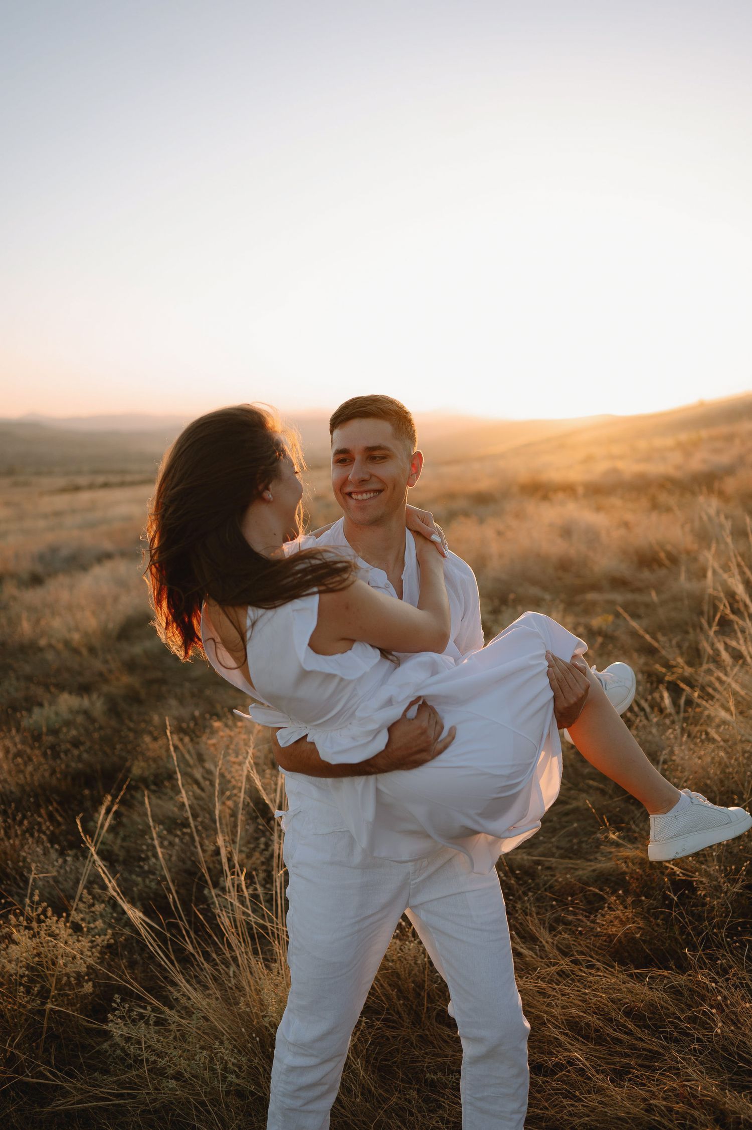 A couple in white outfits share intimate moments while dancing together in a golden field at sunset.