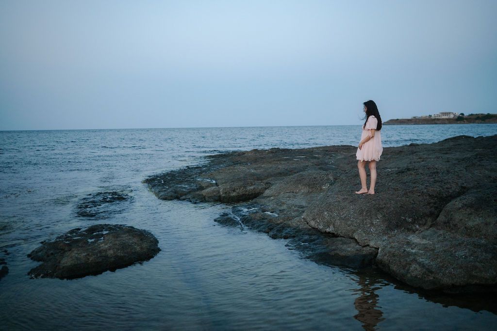 A figure stands on rocky coastal outcrops at sunset overlooking the calm ocean waters.