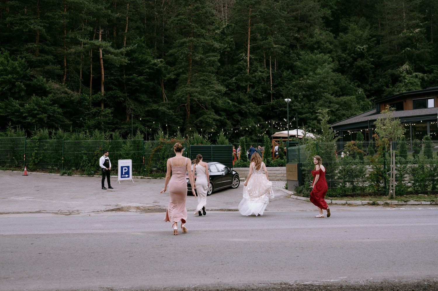 People in formal attire gather in a parking lot area with trees and forest in the background.