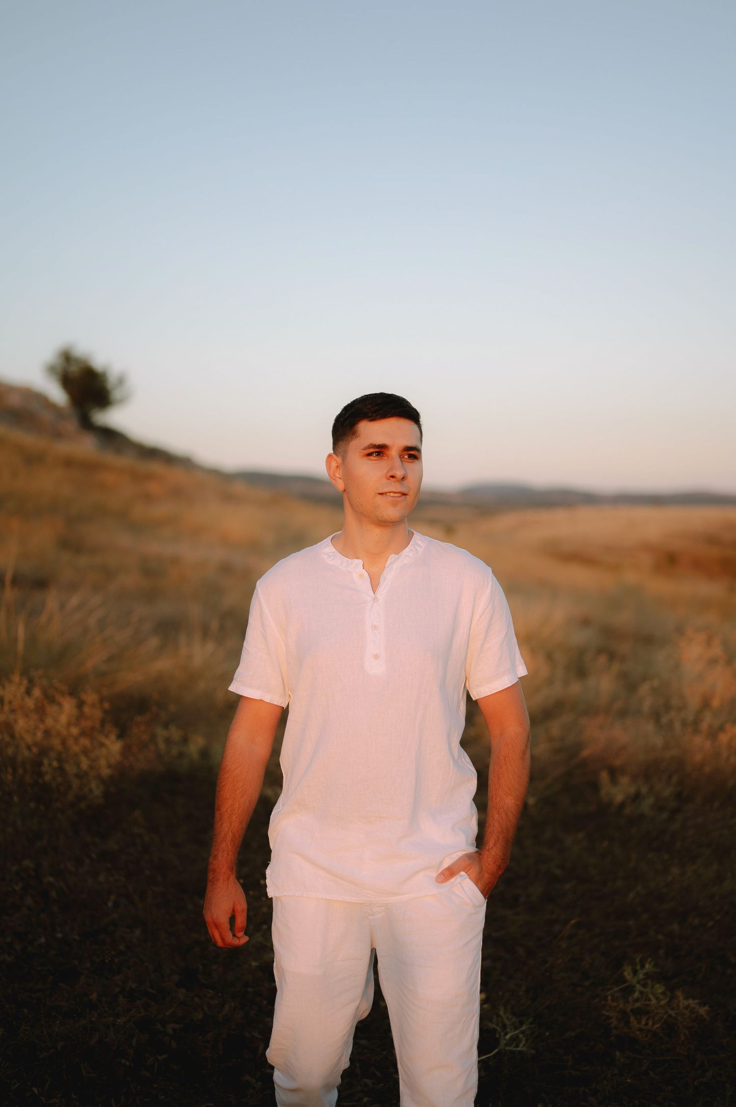 A person in white clothing stands in a golden field at sunset with mountains in the background.
