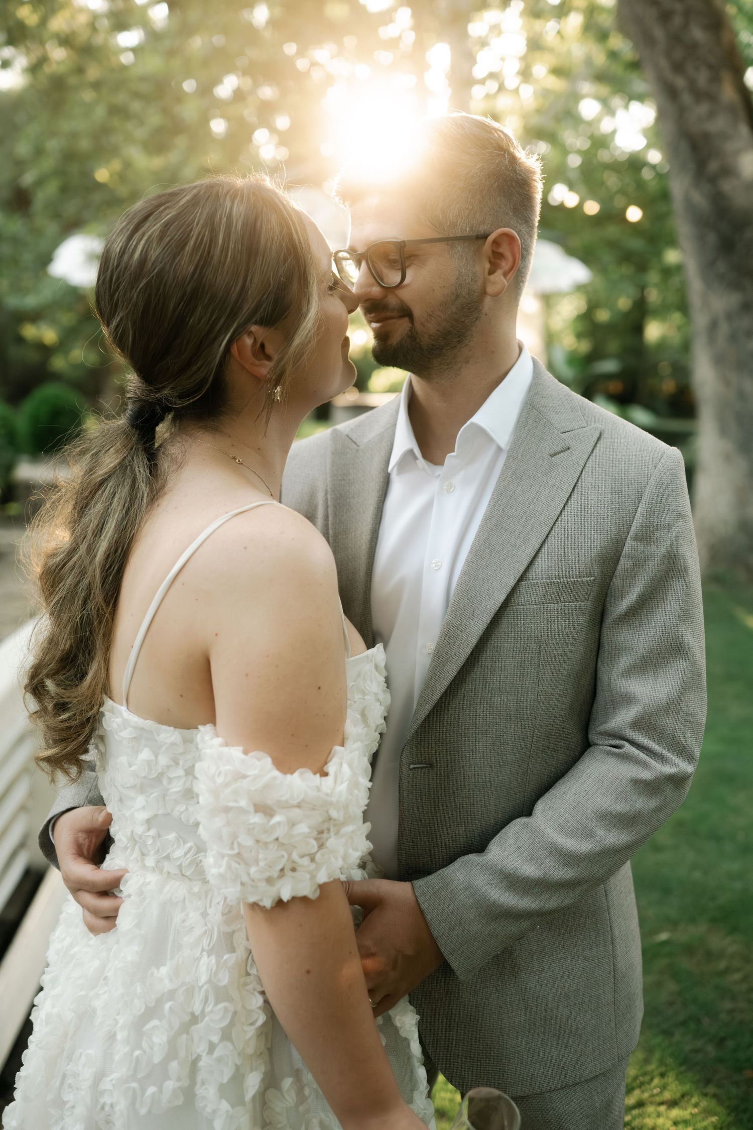 A romantic wedding couple shares tender moments in an outdoor garden setting with soft sunlight filtering through trees.
