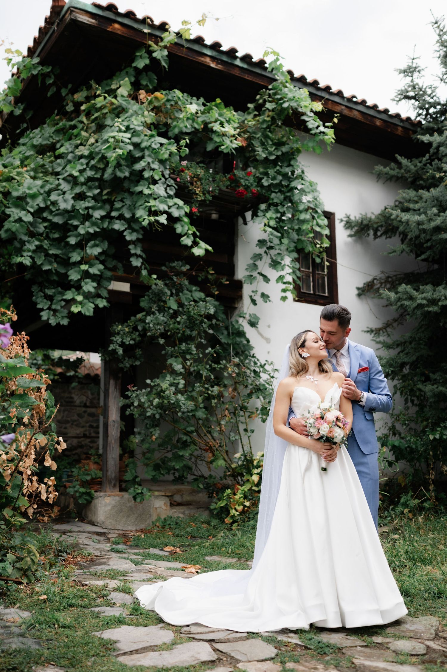 A couple shares a romantic moment in their wedding attire against a white wall covered in lush green vines and foliage.