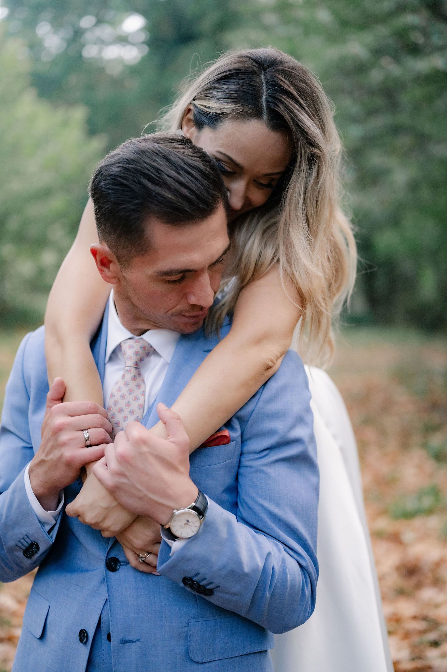 Romantic couple embraces in outdoor setting with groom wearing light blue suit and bride in white dress.