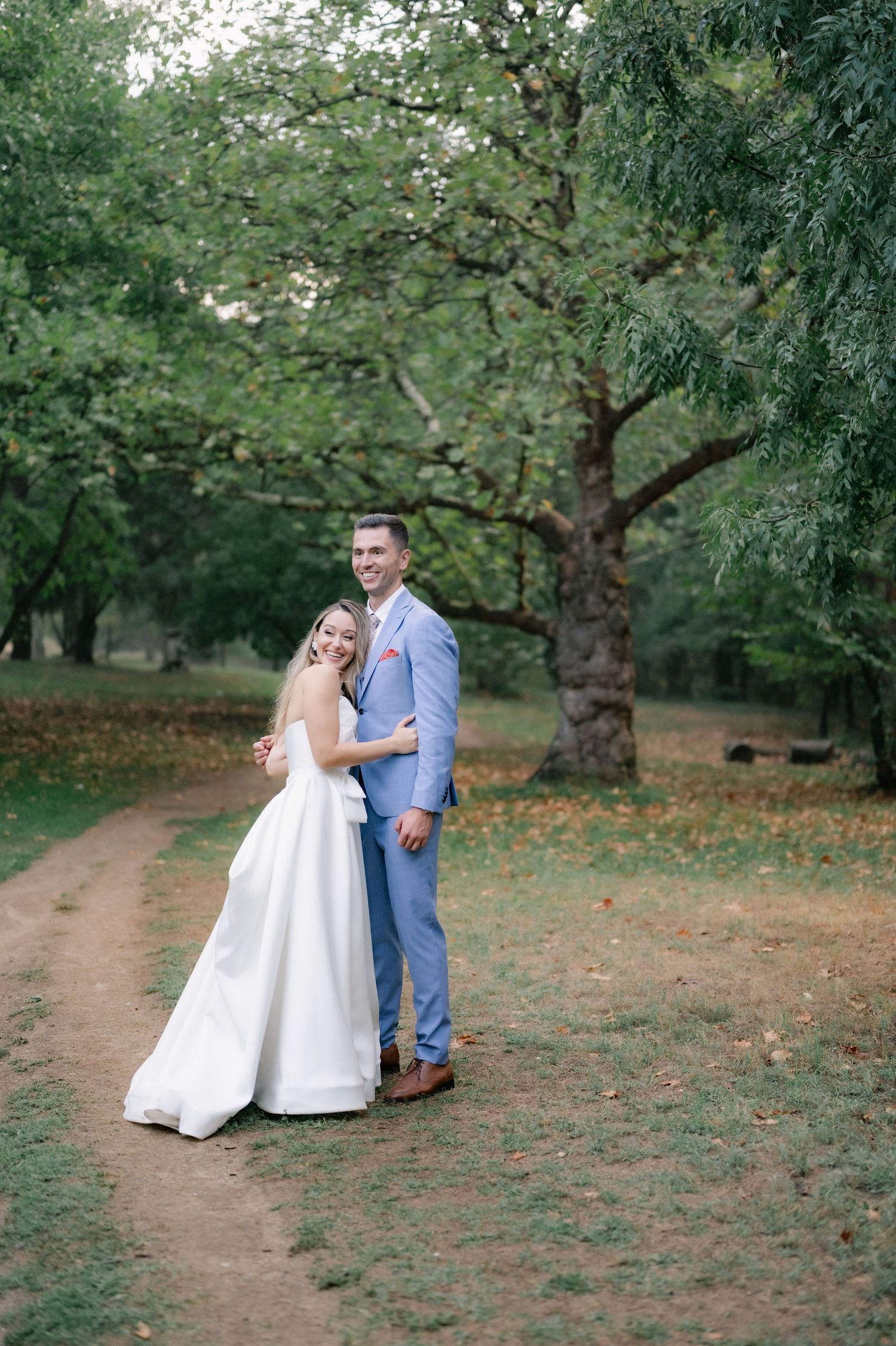 A couple in wedding attire poses on a dirt path surrounded by lush green trees in a park setting.