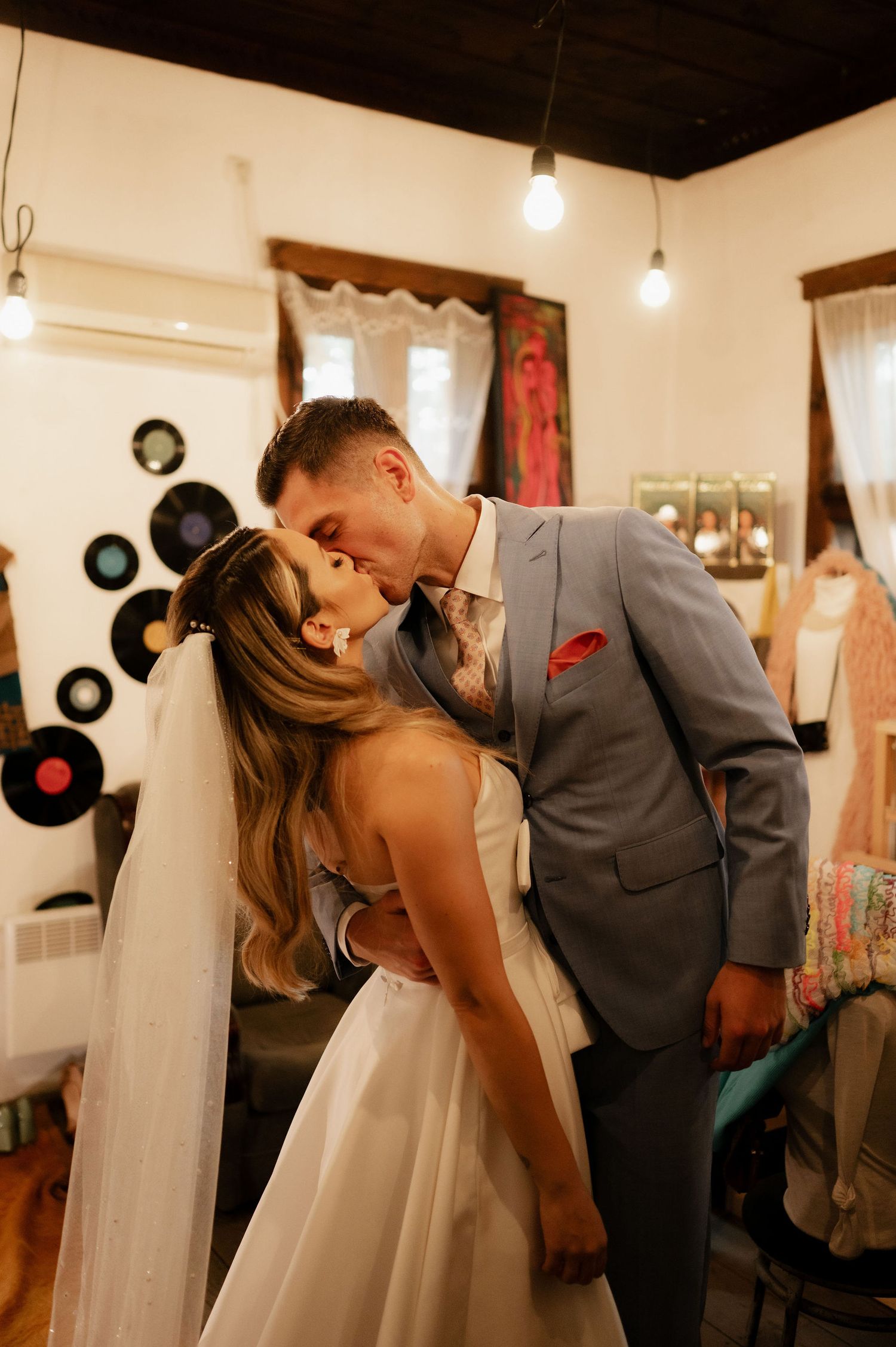 A romantic wedding moment captured in a vintage-styled room decorated with vinyl records on the wall and hanging lights.